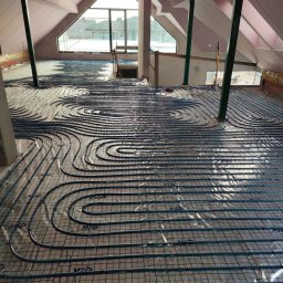 SANITARKA Marcin Skutnik - Instalacja ogrzewania podłogowego w jasnym poddaszu z oknem. Rury ułożone na siatce, widoczne elementy konstrukcji dachu i pionowe podpory.