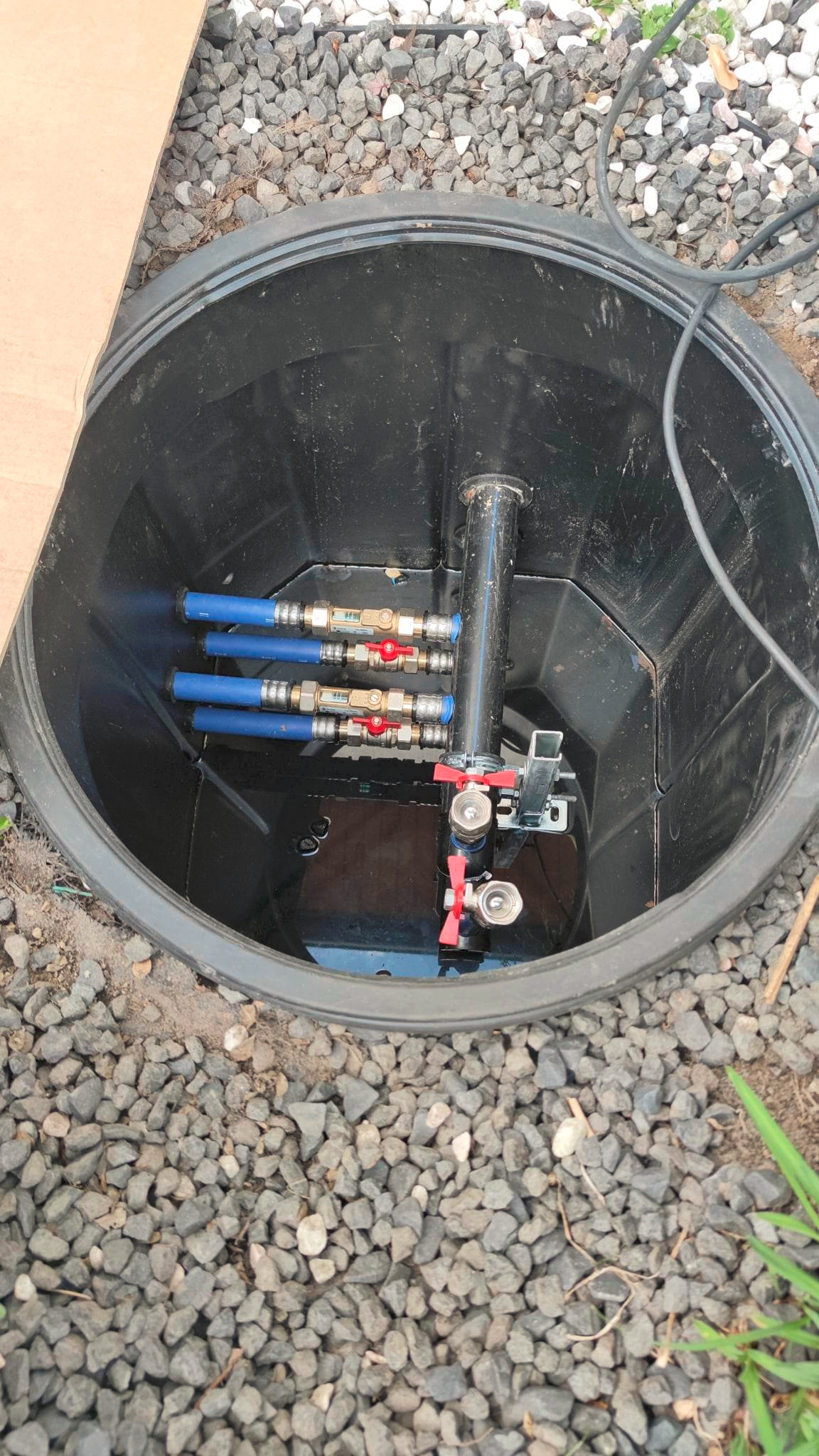 Studzienka rewizyjna z zainstalowanymi zaworami i rurami, otoczona żwirem. Widoczne elementy instalacji wodnej wewnątrz czarnej studzienki.