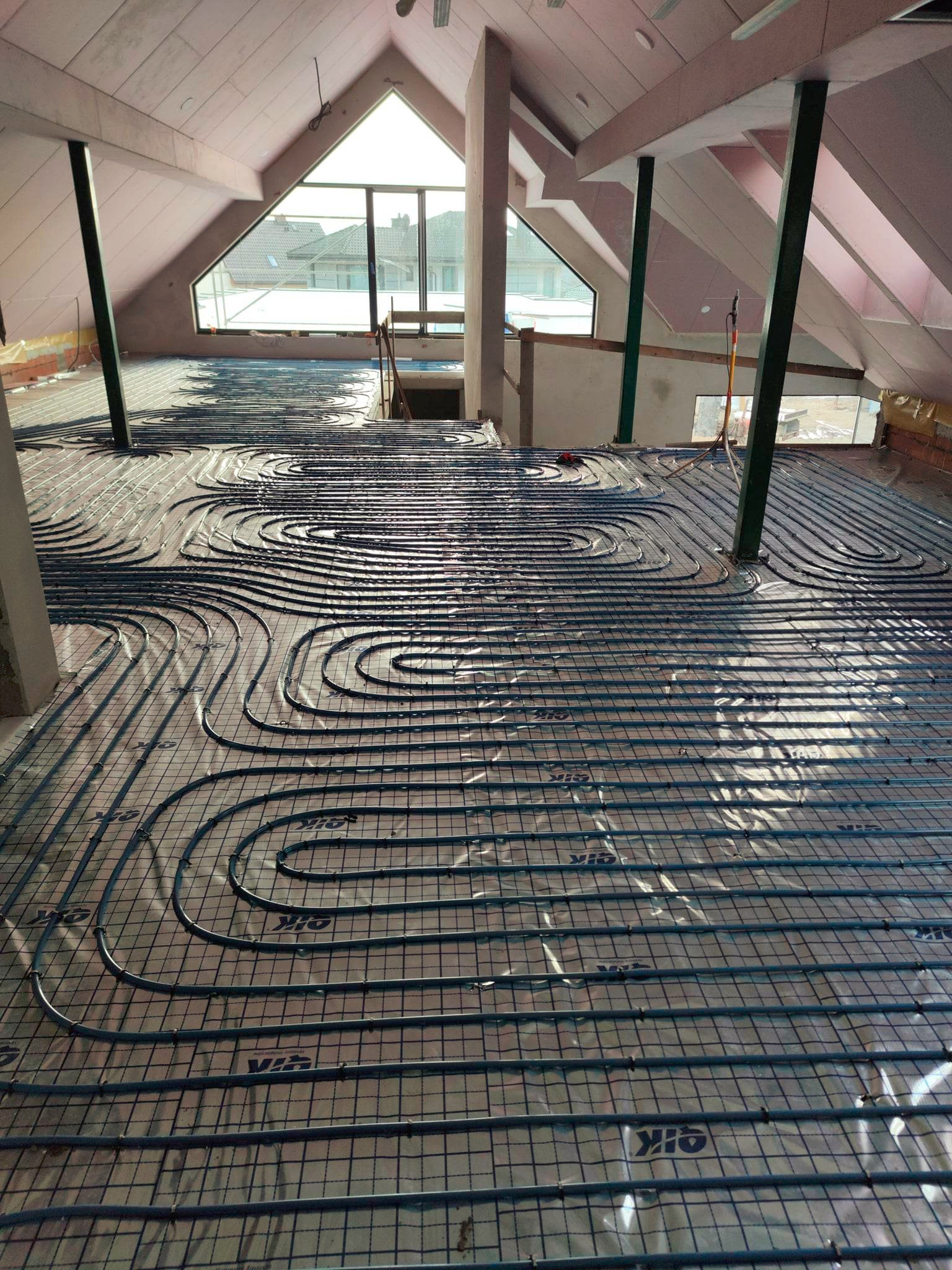 Instalacja ogrzewania podłogowego w jasnym poddaszu z oknem. Rury ułożone na siatce, widoczne elementy konstrukcji dachu i pionowe podpory.