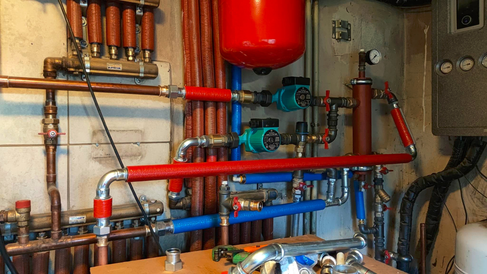 Złożona instalacja grzewcza z czerwonym zbiornikiem wyrównawczym, pompami obiegowymi i rurami miedzianymi oraz niebieskimi, częściowo zaizolowanymi, w kotłowni.