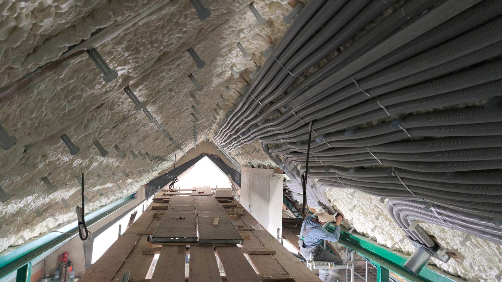 Izolacja poddasza pianą PUR i równolegle ułożone rury wentylacyjne, widok z rusztowania, pracownik w trakcie montażu,  szczegóły mocowania.