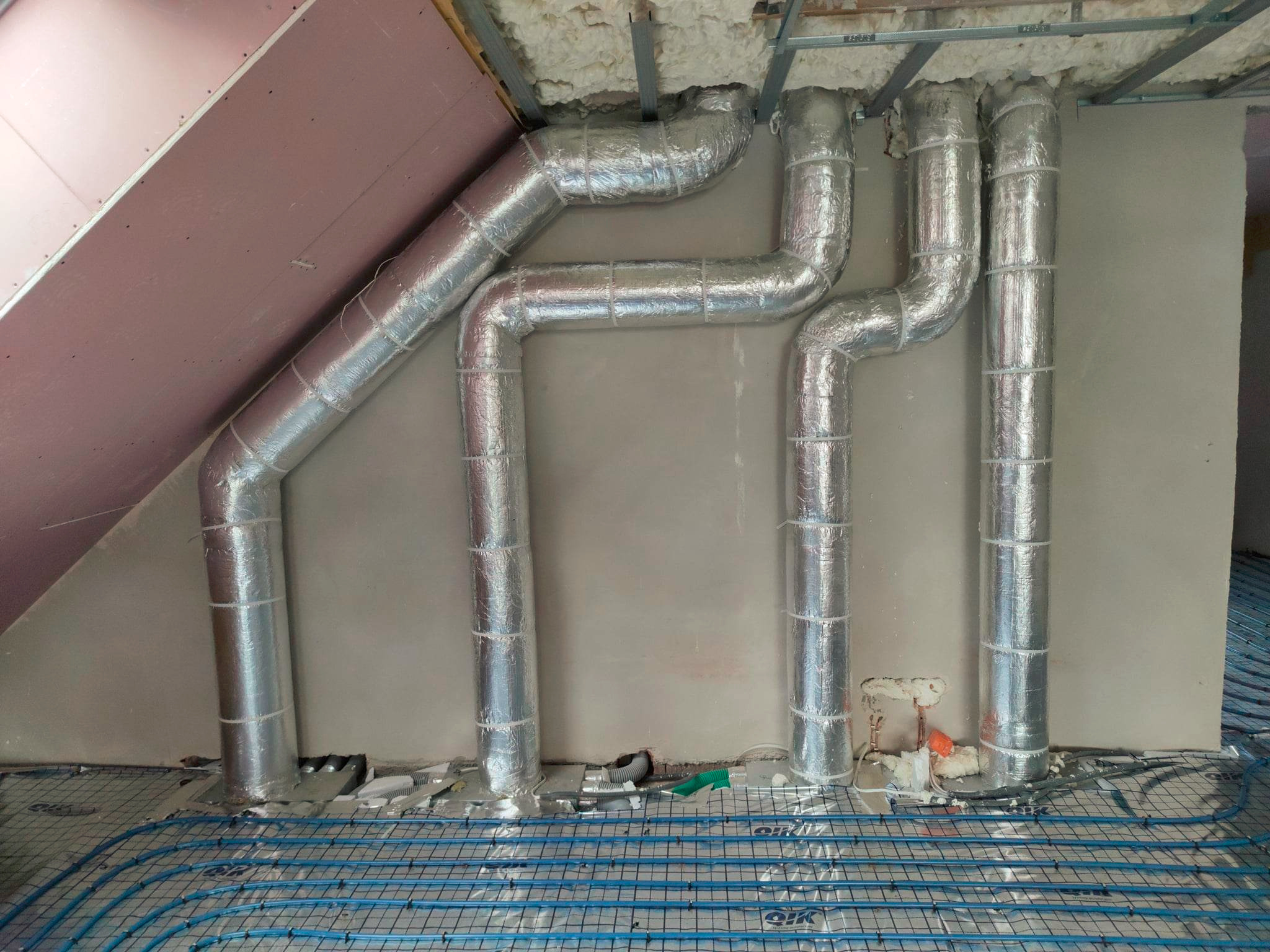 Izolowane rury wentylacyjne z folii aluminiowej przymocowane do sufitu, nad niebieską instalacją ogrzewania podłogowego w trakcie montażu.