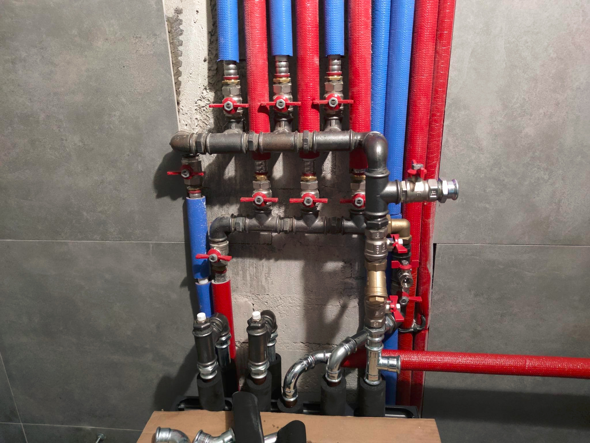 Węzeł instalacji centralnego ogrzewania z rurami w izolacji czerwonej i niebieskiej, zaworami z czerwonymi pokrętłami na tle ściany z szarych płytek.