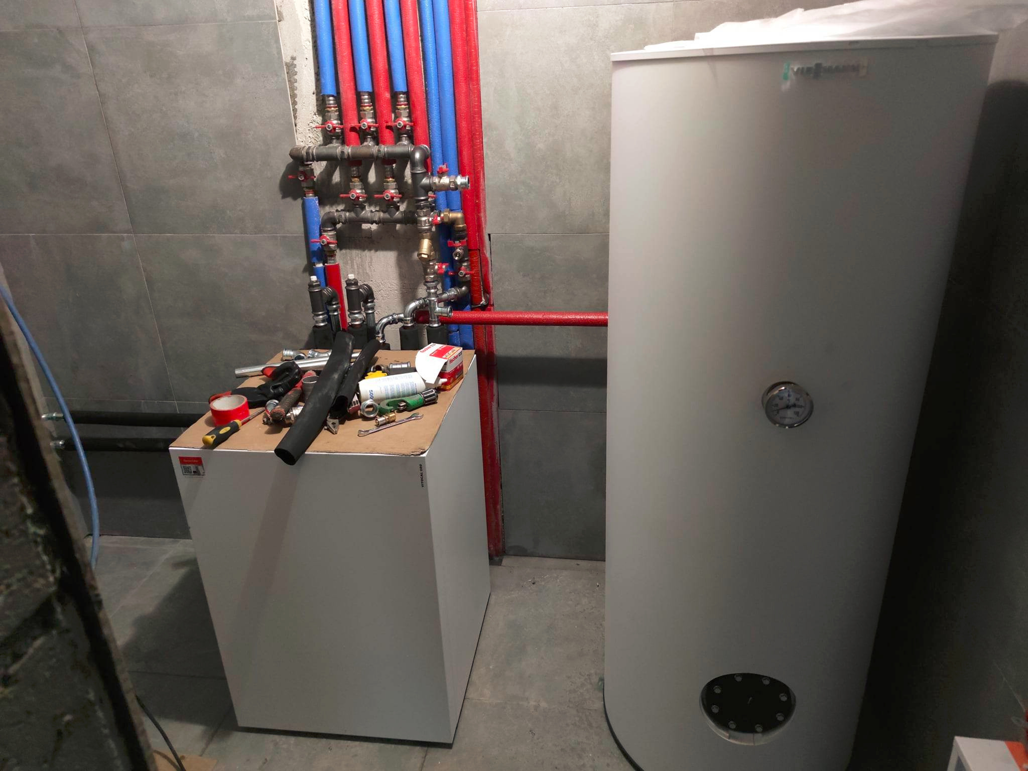 Nowoczesna kotłownia: biały piec Viessmann, instalacja z rur miedzianych i niebieskich, narzędzia na białej obudowie, szare płytki na ścianie.