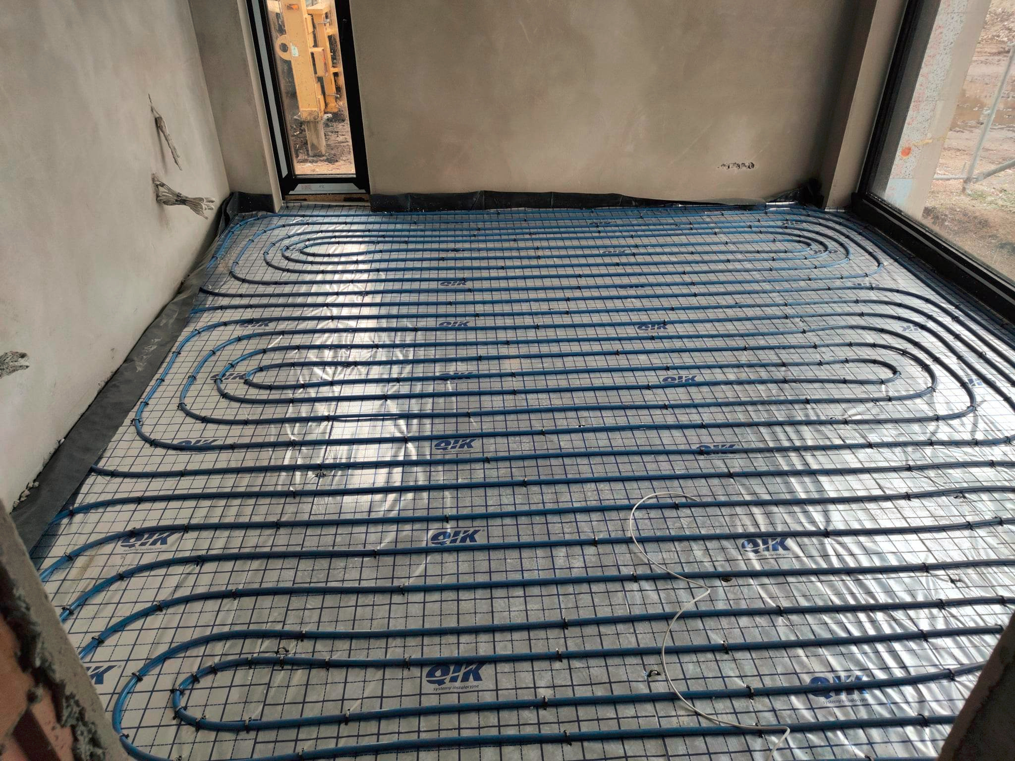 Niebieskie rury ogrzewania podłogowego ułożone na izolacji, widoczne okna i surowa ściana. Etap instalacji w nowym budynku.