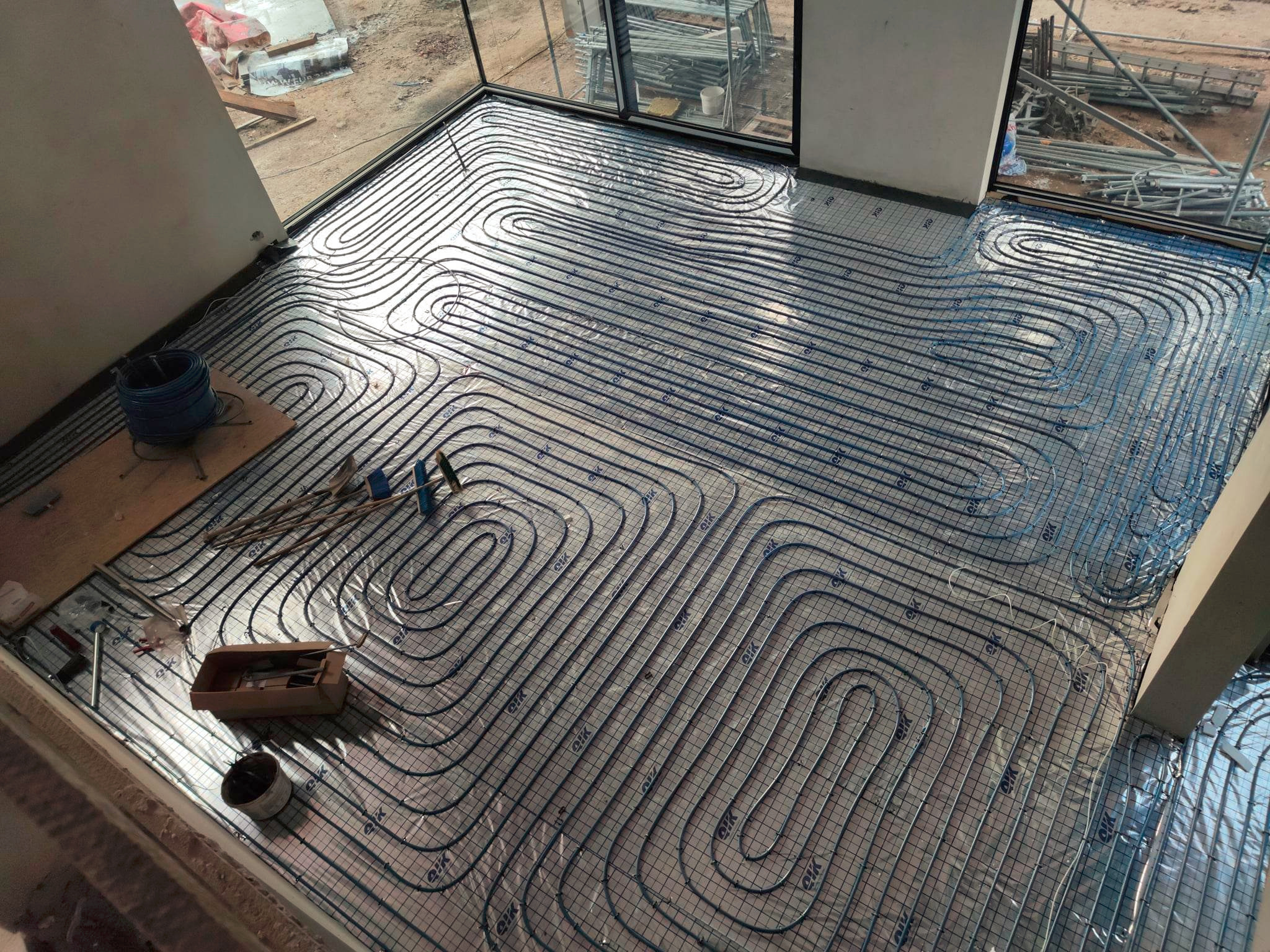 Niebieskie rury ogrzewania podłogowego ułożone na folii izolacyjnej z siatką, widoczne narzędzia i elementy montażowe. Duże okna w tle.