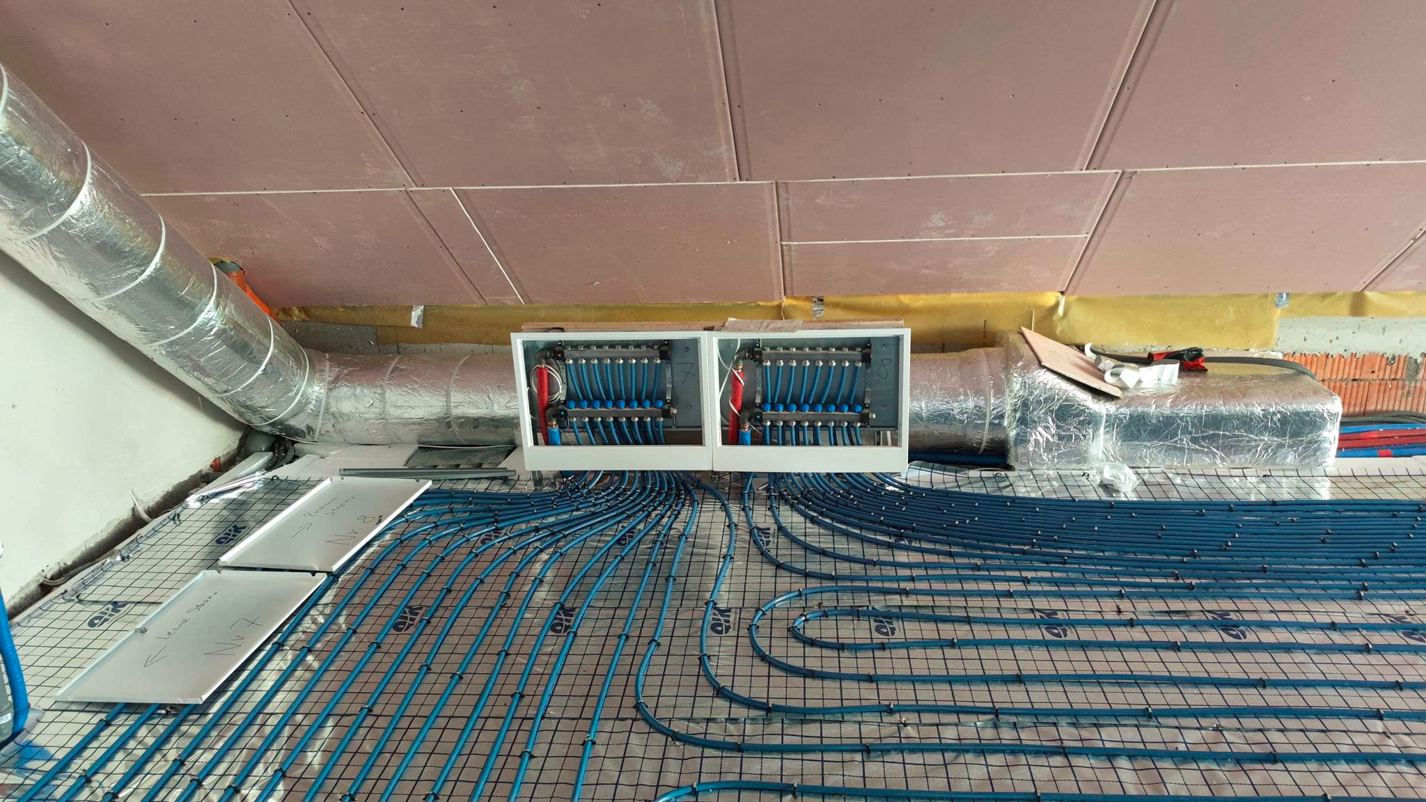 Niebieskie rury ogrzewania podłogowego ułożone na izolacji z rozdzielaczem w białej obudowie, widok z góry na poddaszu w trakcie instalacji.