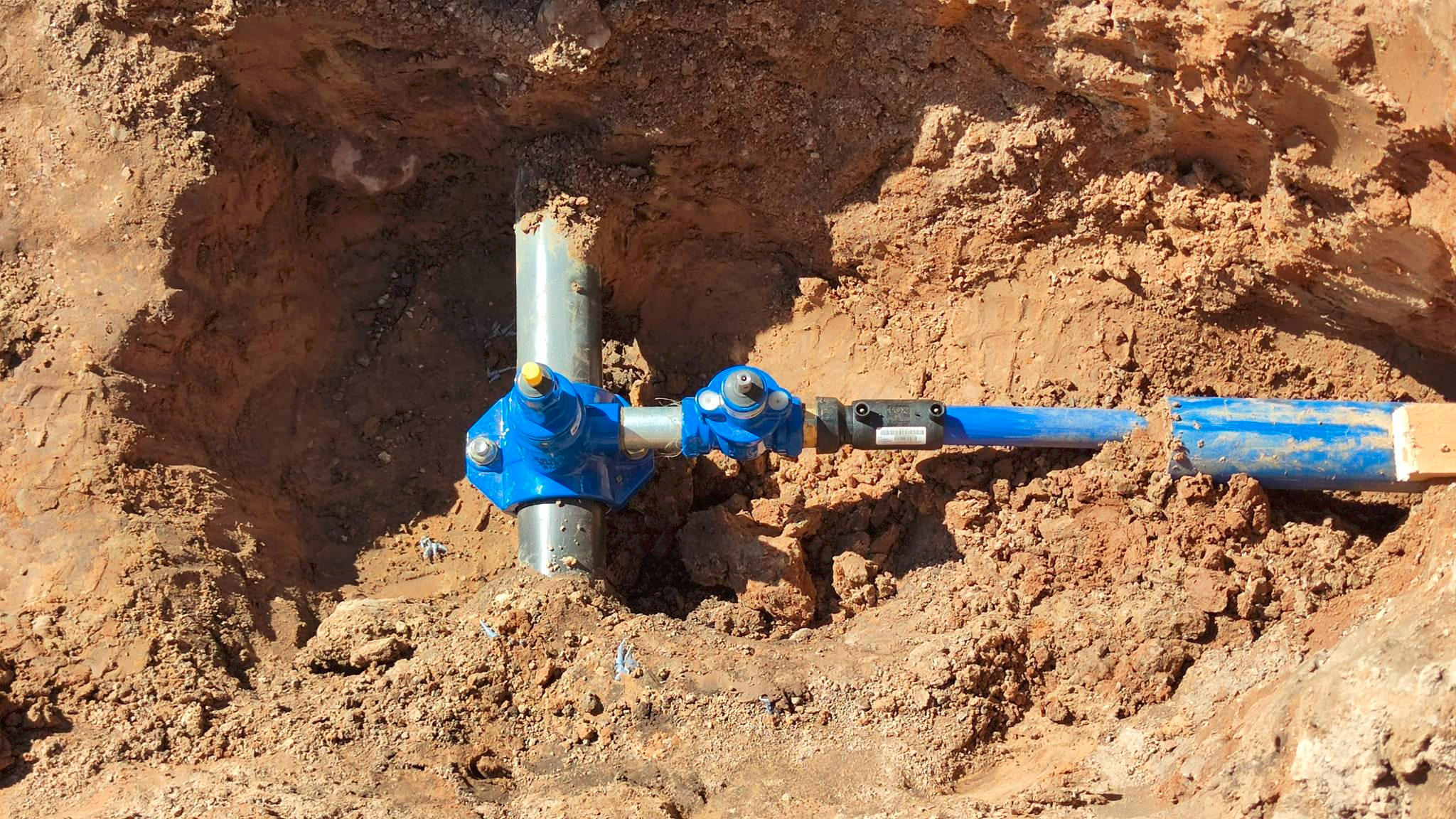 Niebieskie złącze instalacji wodnej w wykopie ziemnym, widoczne rury z tworzywa i metalu, żółte elementy regulacyjne. Ujęcie z góry.