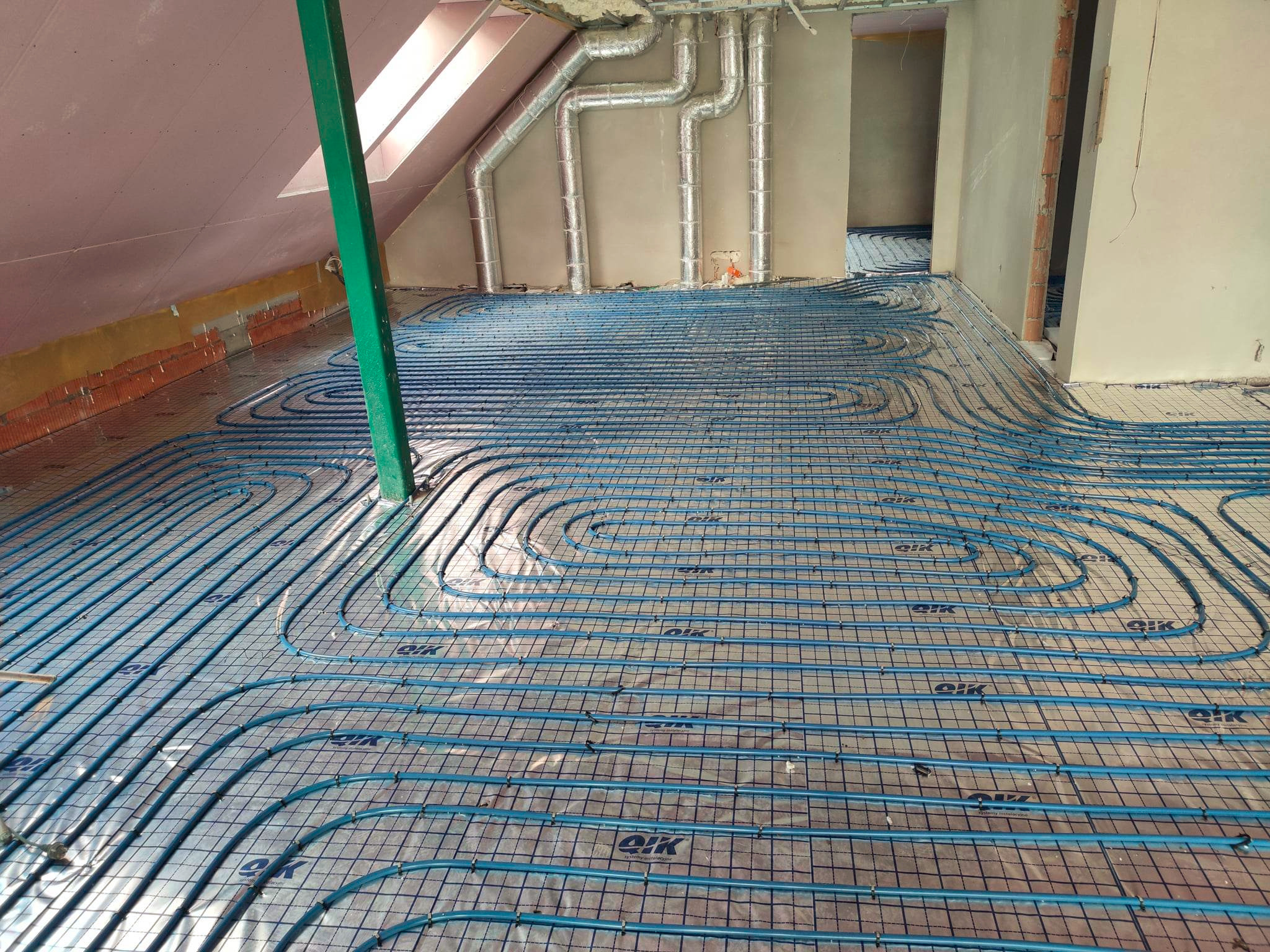 Niebieskie rury ogrzewania podłogowego ułożone na folii w nowym budynku z widocznymi przewodami wentylacyjnymi na suficie i oknem dachowym.