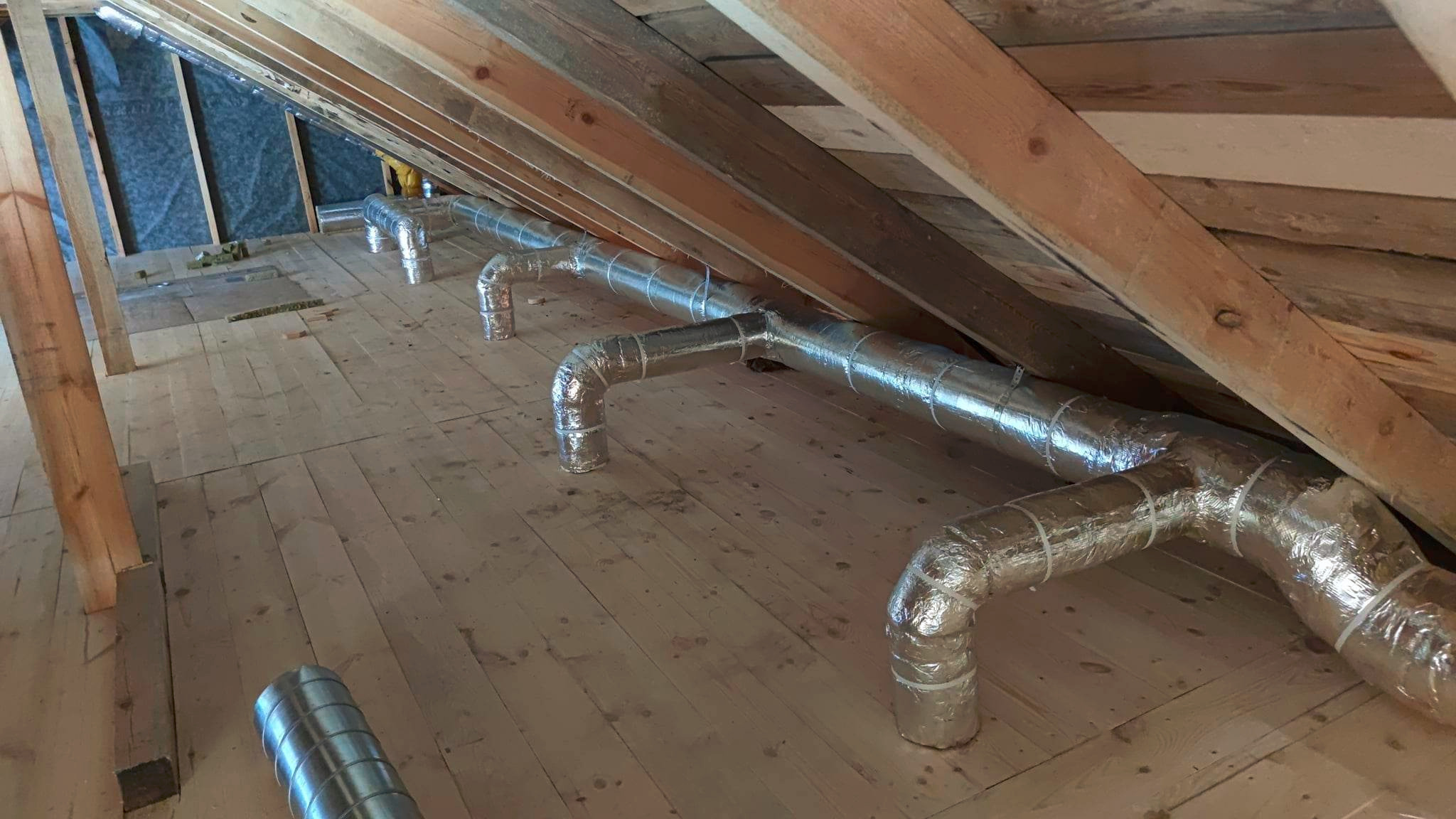 Instalacja wentylacyjna z izolowanych rur aluminiowych w przestrzeni poddasza, widoczne drewniane elementy konstrukcyjne dachu i podłogi.