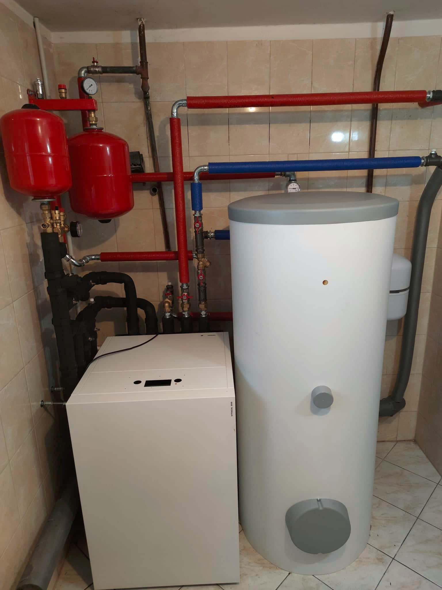 Nowoczesny piec CO Viessmann obok białego zasobnika ciepłej wody, z widocznymi czerwonymi zbiornikami wyrównawczymi i instalacją w tle, na tle beżowych płytek.