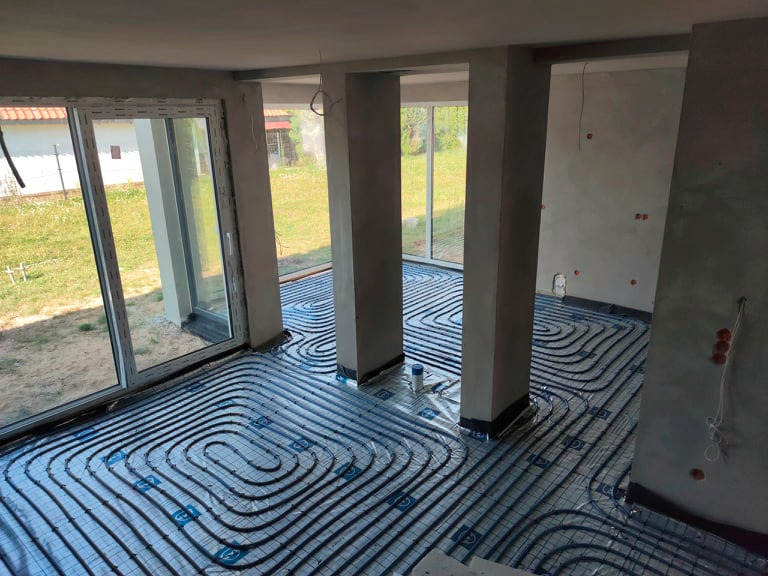 Szczegółowe ułożenie rur ogrzewania podłogowego na folii, w surowym wnętrzu z kolumnami i dużym oknem tarasowym. Widoczne elementy instalacji elektrycznej na ścianie.