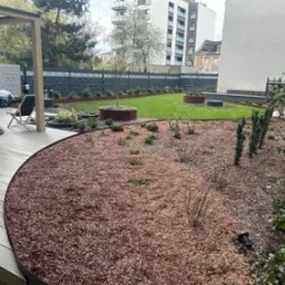 Nowoczesny ogród z trawnikiem, rabatami wysypanymi korą i donicami z roślinami, widoczny fragment tarasu z drewnianą podłogą i wiszącym fotelem, w tle budynki mieszkalne.