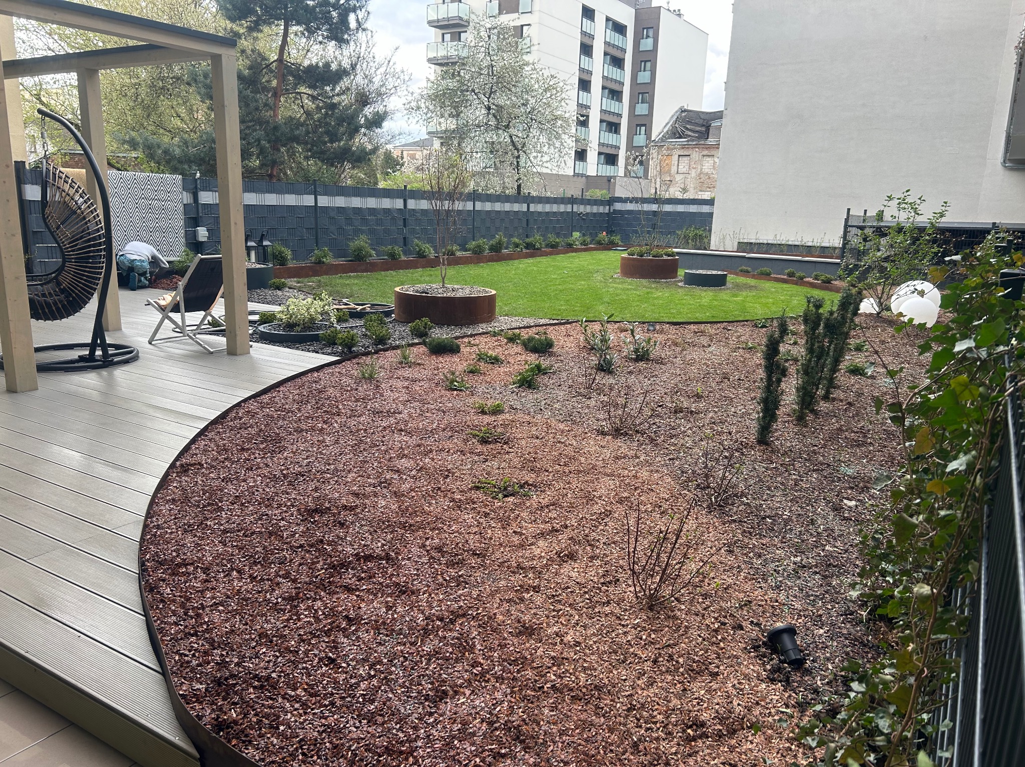 Nowoczesny ogród z trawnikiem, rabatami wysypanymi korą i donicami z roślinami, widoczny fragment tarasu z drewnianą podłogą i wiszącym fotelem, w tle budynki mieszkalne.