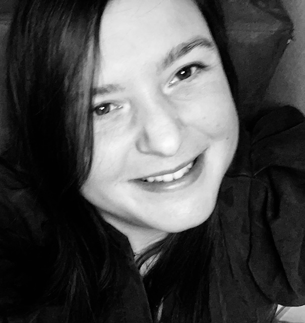 Uśmiechnięta brunetka w czerni, portret w odcieniach szarości. Widoczne włosy opadające na ramiona i fragment ciemnego tła. Naturalny uśmiech i spojrzenie w obiektyw.