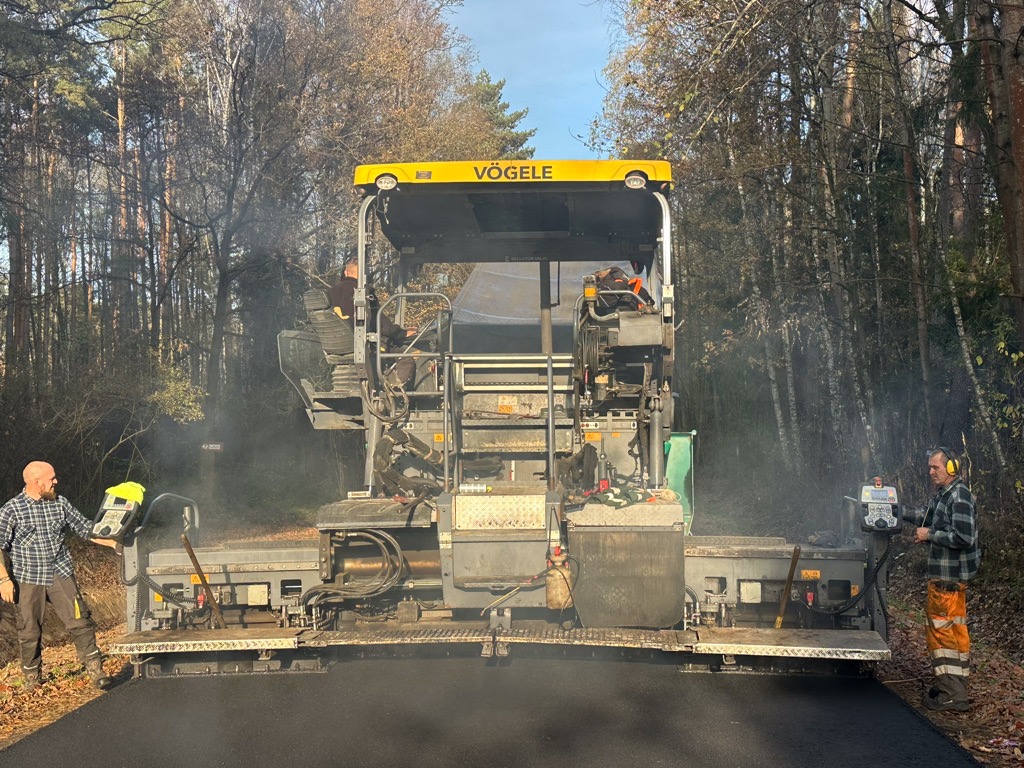 Asfaltowanie drogi leśnej przy użyciu rozściełacza Vögele; dwóch operatorów kontroluje proces, widoczny lekki dym unoszący się znad świeżo położonej nawierzchni.