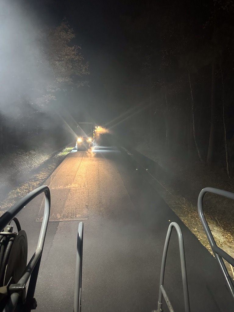 Asfaltowanie drogi nocą, widok z maszyny rozkładającej asfalt, z efektem zamglenia i światłami pojazdów w tle.