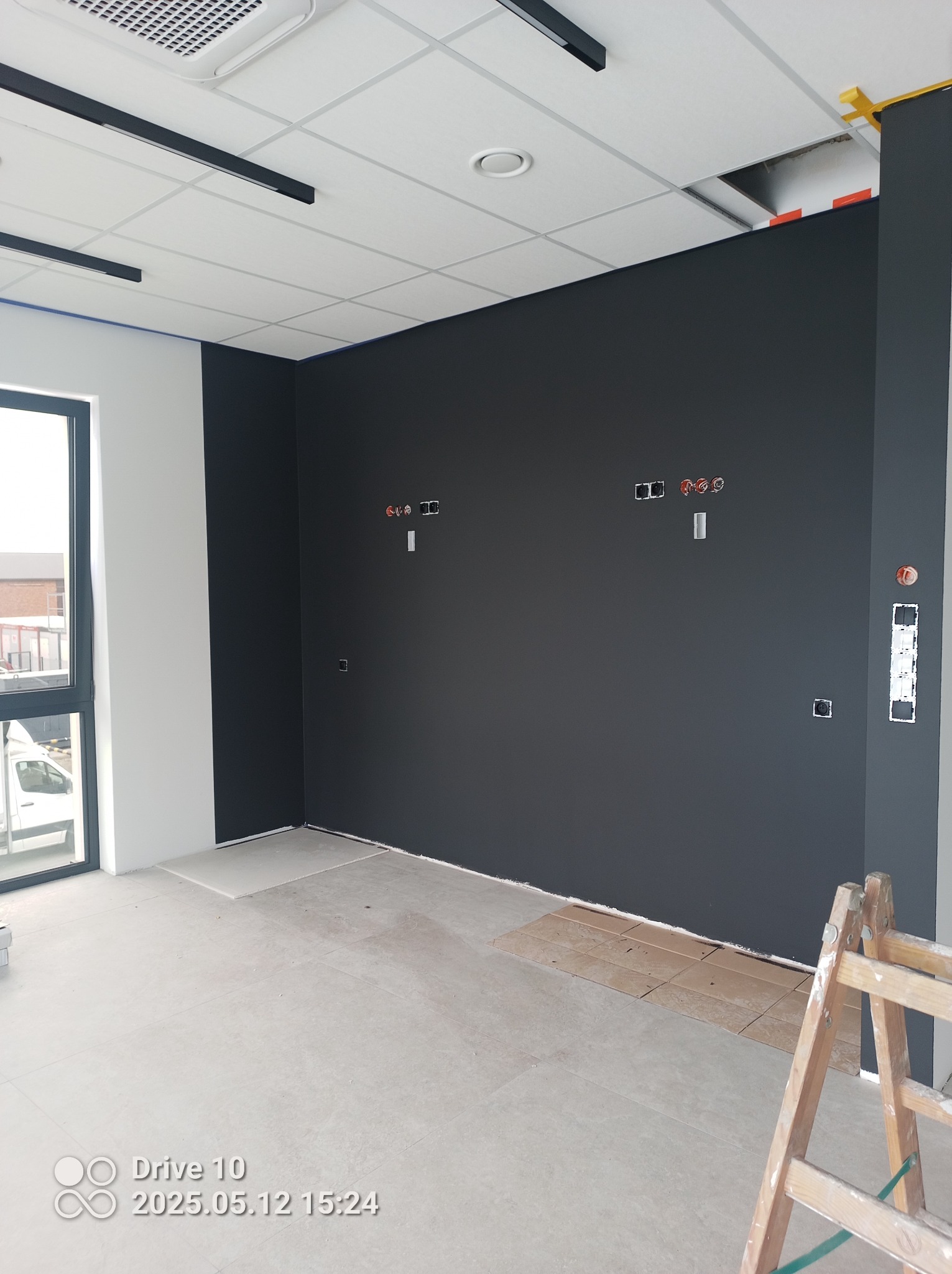 Ściana pomalowana na ciemny kolor z widocznymi puszkami elektrycznymi we Włodzimierzowie. Fragment podłogi zabezpieczony kartonem, drabina malarska w rogu.