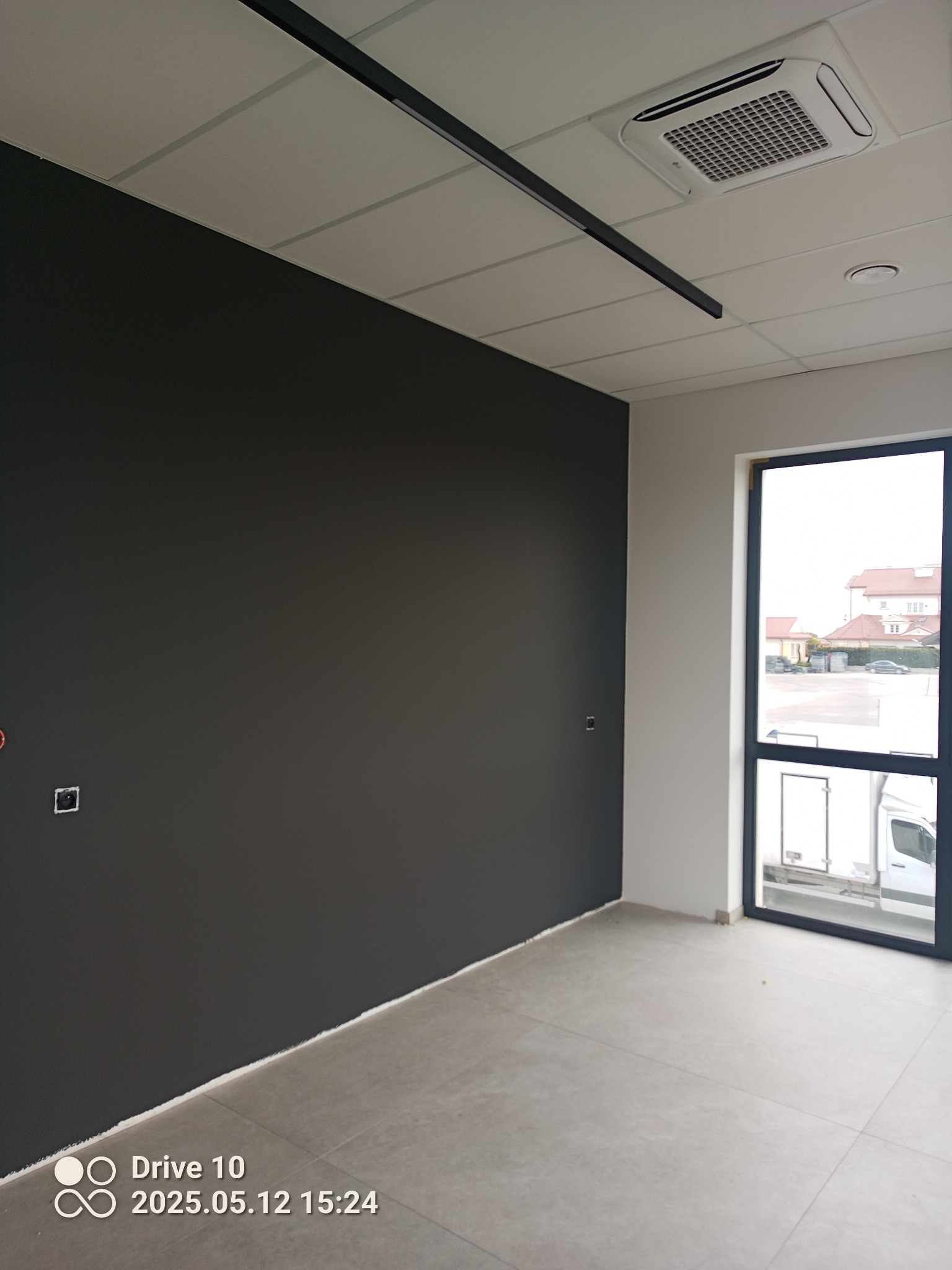 Świeżo pomalowana ściana w ciemnym kolorze, jasna podłoga i sufit z klimatyzacją. W oknie widoczny budynek i samochody. Minimalistyczny styl wnętrza.