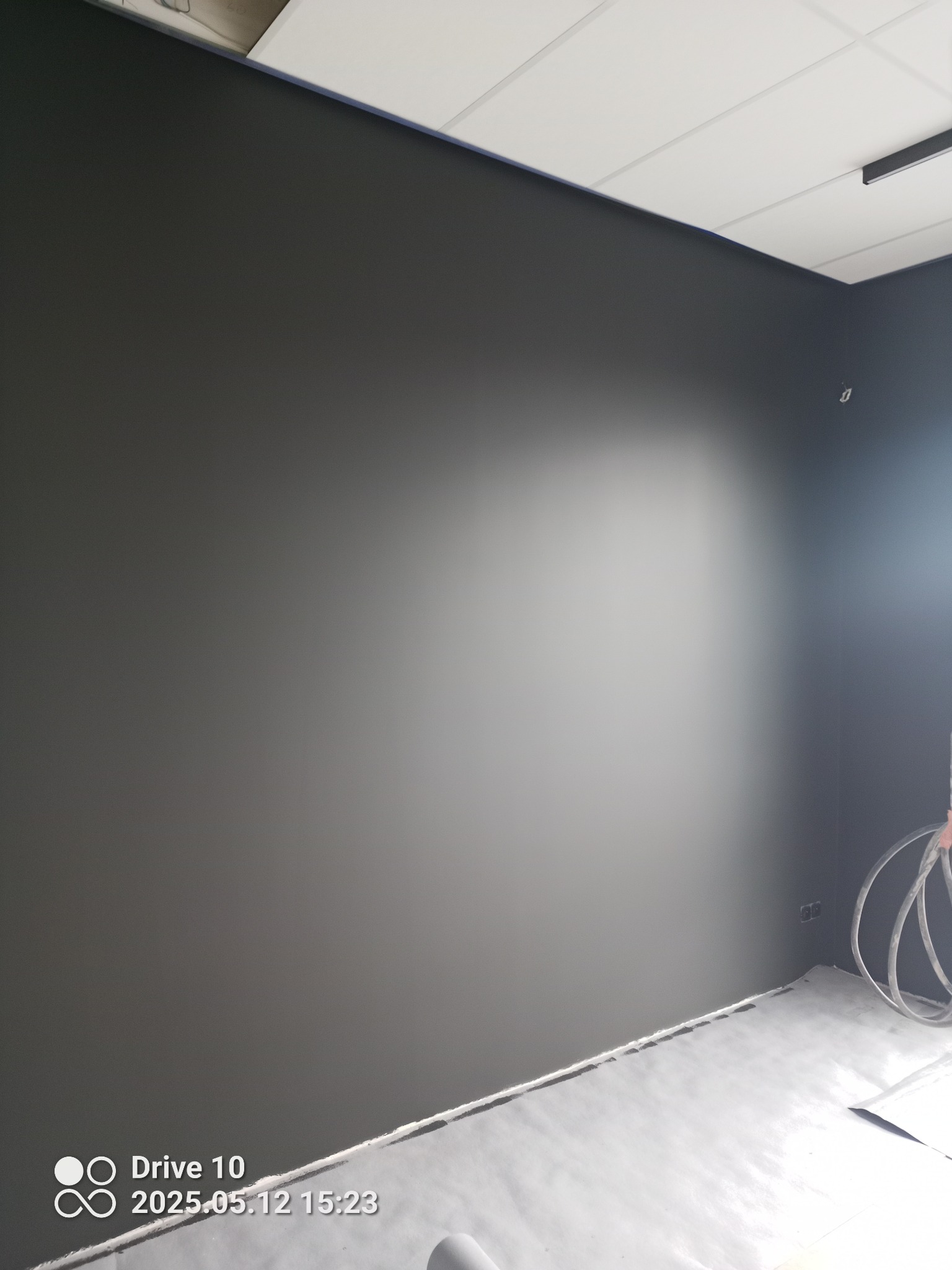 Świeżo pomalowana ściana w odcieniach szarości w nowym wnętrzu, z widocznymi gniazdkami elektrycznymi i zabezpieczoną podłogą folią malarską.