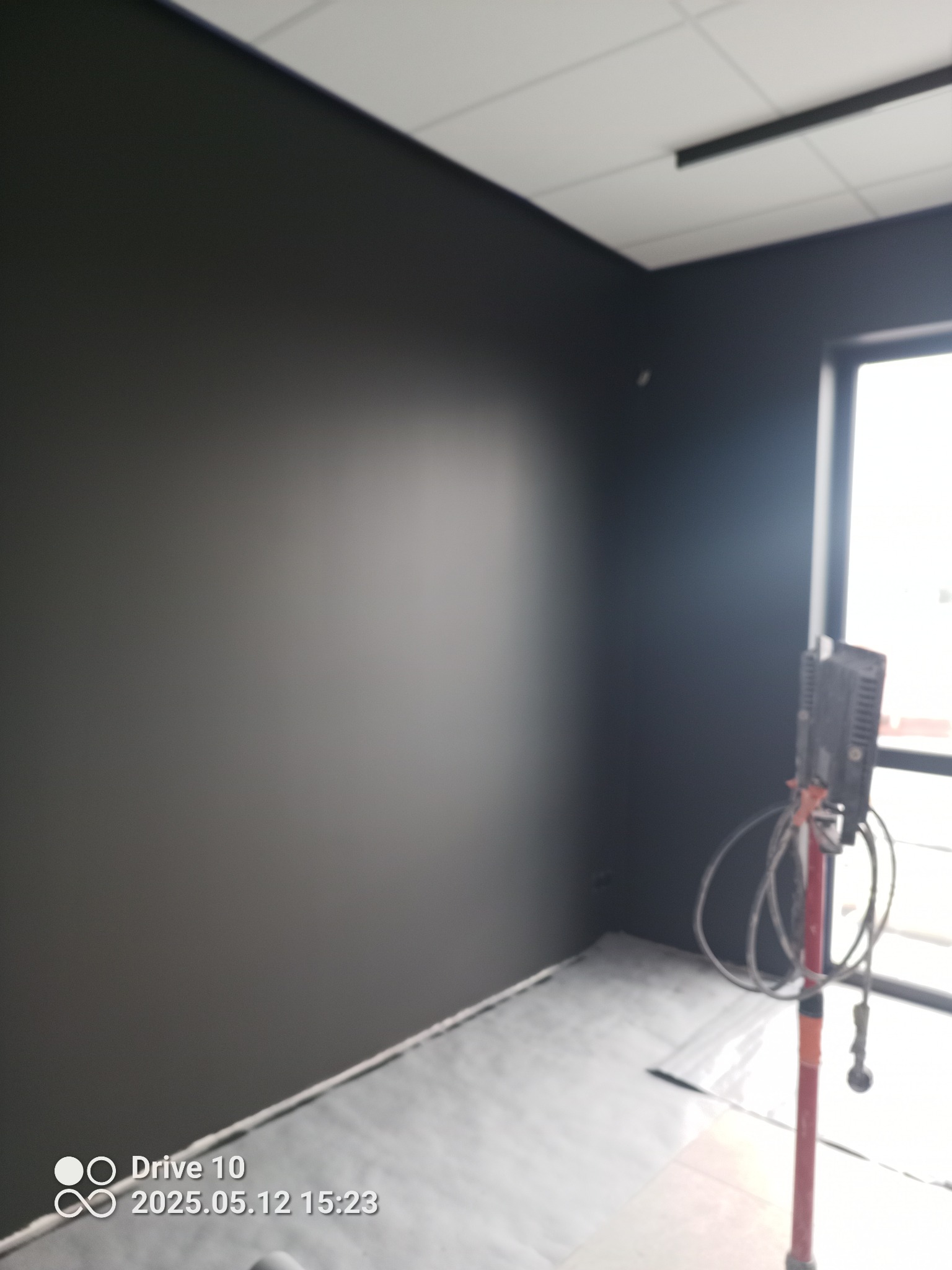 Pomieszczenie po malowaniu natryskowym na ciemny kolor. Widoczny sprzęt do malowania i zabezpieczona podłoga. Ściany matowe, jednolite.