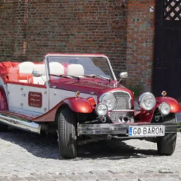 Retro, czerwono-biały kabriolet z tablicą 'GO BARON' na brukowanym podłożu, obok ceglanej ściany i drewnianej bramy. Widoczna tabliczka z numerem telefonu.