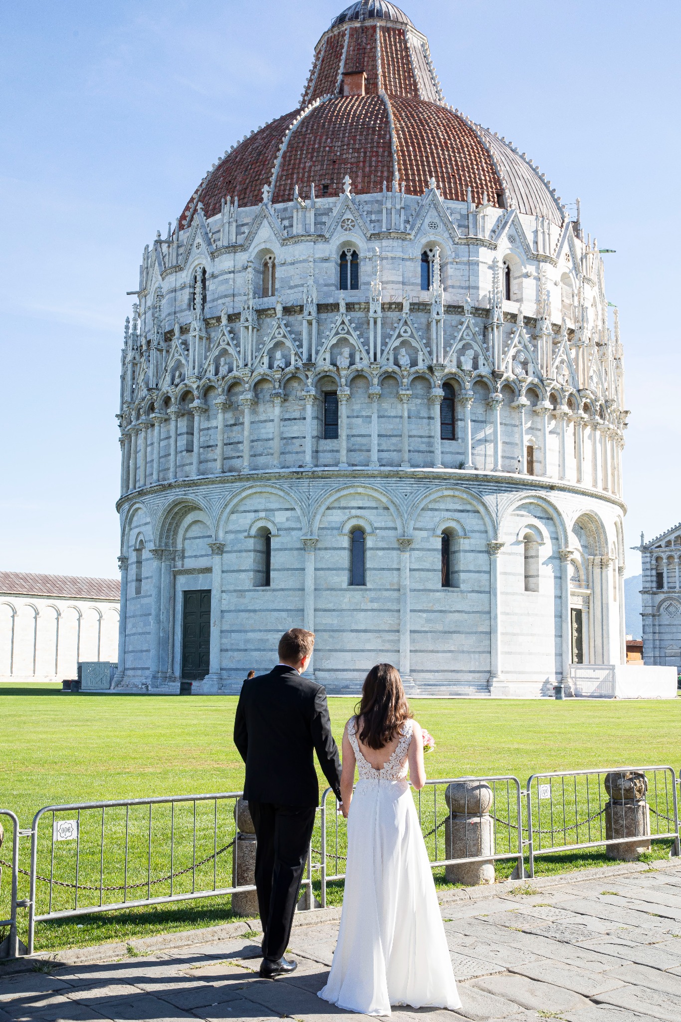 Para młoda, tyłem, trzymająca się za ręce, na tle Baptysterium w Pizie w słoneczny dzień. Suknia panny młodej z koronkowymi detalami. Elegancki garnitur pana młodego.