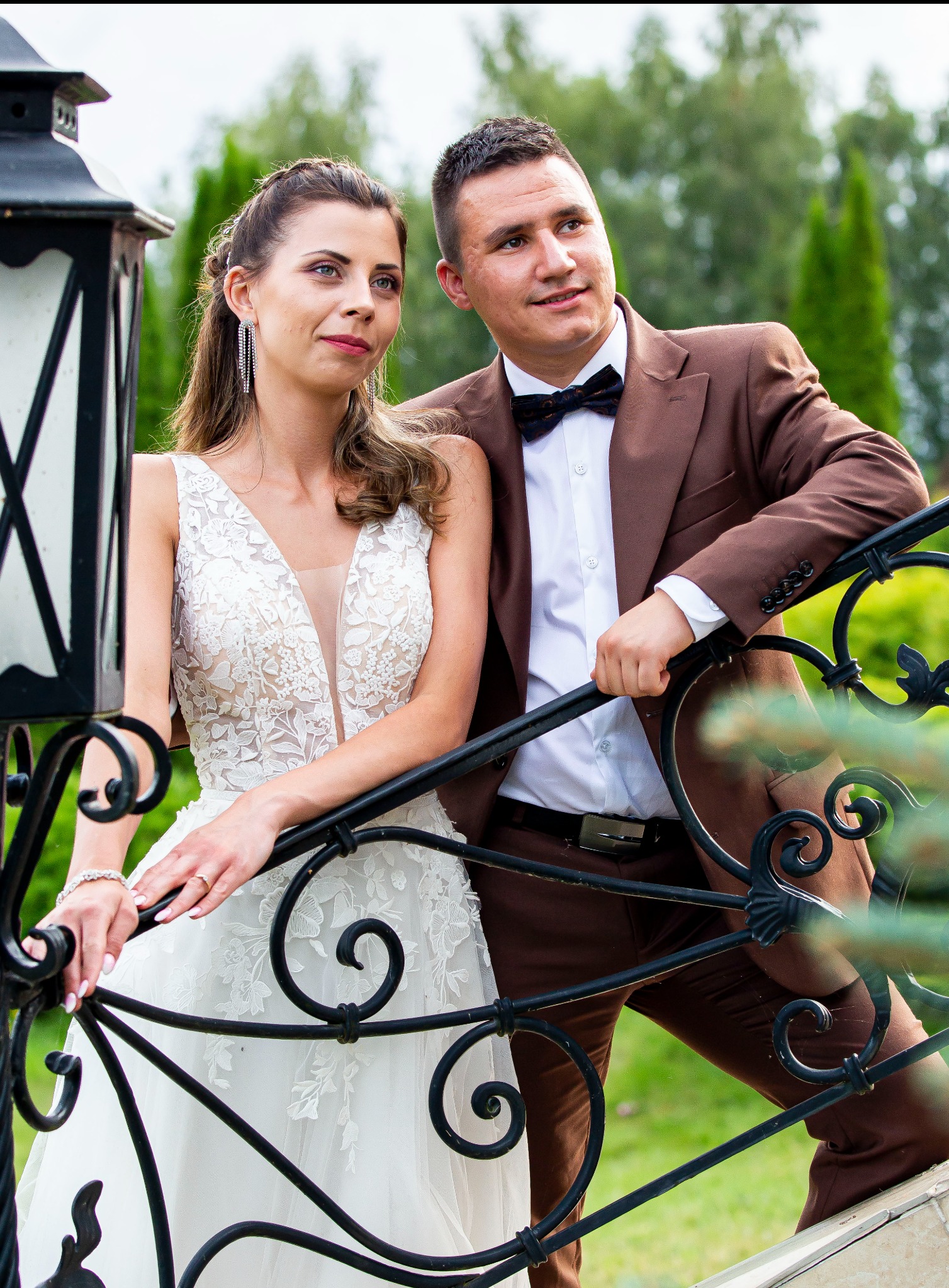 Elegancka para młoda w brązowym garniturze i koronkowej sukni pozuje przy ozdobnej, czarnej balustradzie w plenerze. Romantyczna sesja ślubna.