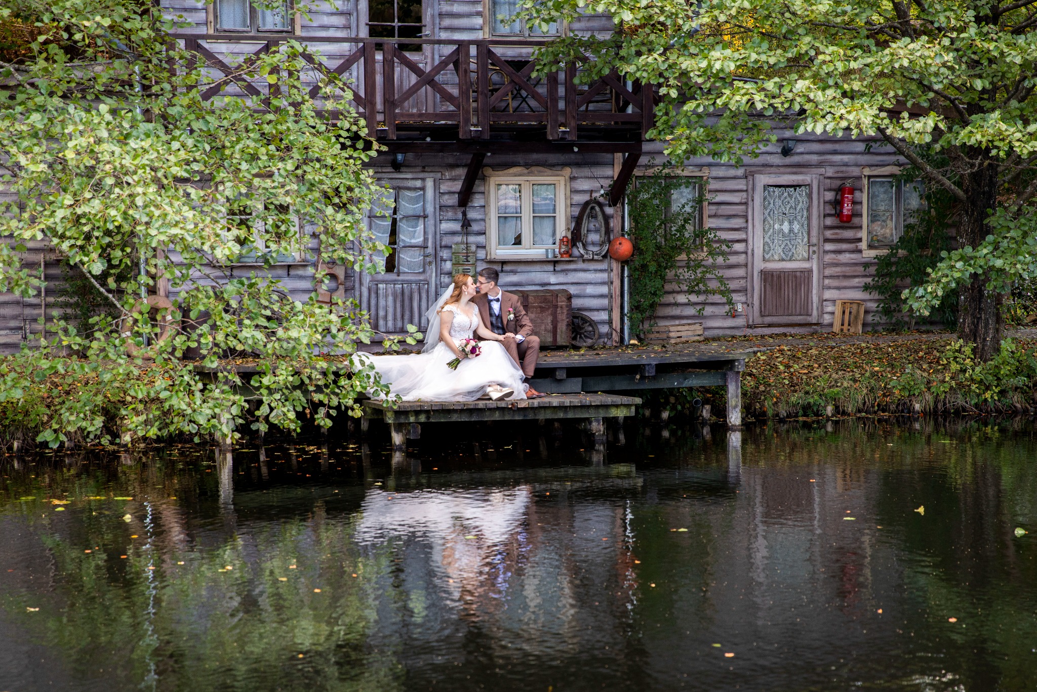 Para młoda siedzi na pomoście nad wodą, całując się na tle drewnianego domku z balkonem, w otoczeniu zieleni i refleksów wody. Romantyczna sceneria.