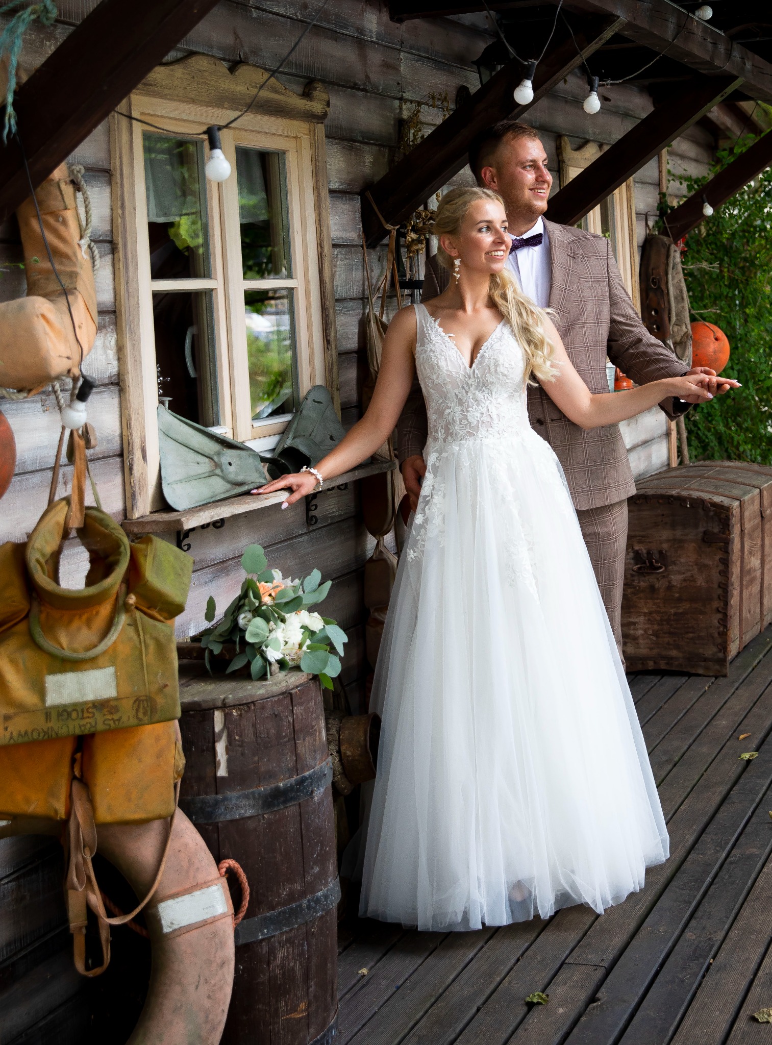 Uśmiechnięta para młoda w plenerze: panna młoda w koronkowej sukni, pan młody w brązowym garniturze. W tle drewniana chata z marynistycznymi akcentami.