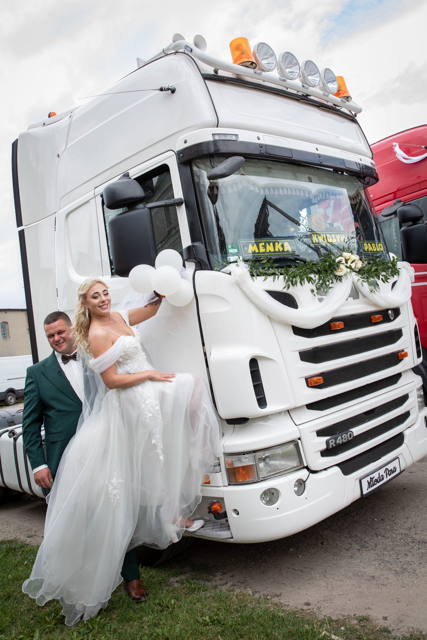 Para młoda pozuje przy białej ciężarówce Scania R480 udekorowanej kwiatami i wstążkami. Panna młoda trzyma białe balony, obok stoi pan młody w zielonym garniturze.