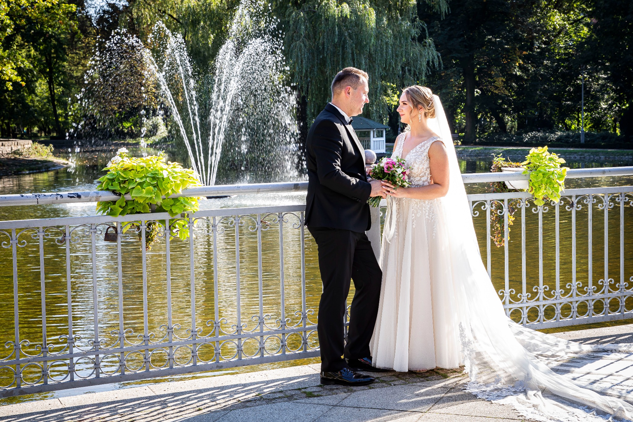 Para młoda w eleganckich strojach stoi na tle fontanny i ozdobnej balustrady z kwiatami w parku, w słoneczny dzień. Romantyczna sesja ślubna.