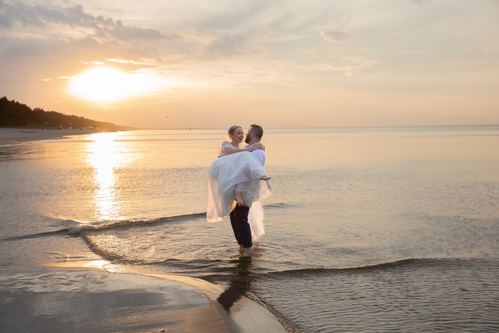 Mężczyzna trzyma pannę młodą na rękach w płytkiej wodzie na plaży o zachodzie słońca. Romantyczna fotografia ślubna z ciepłym światłem.