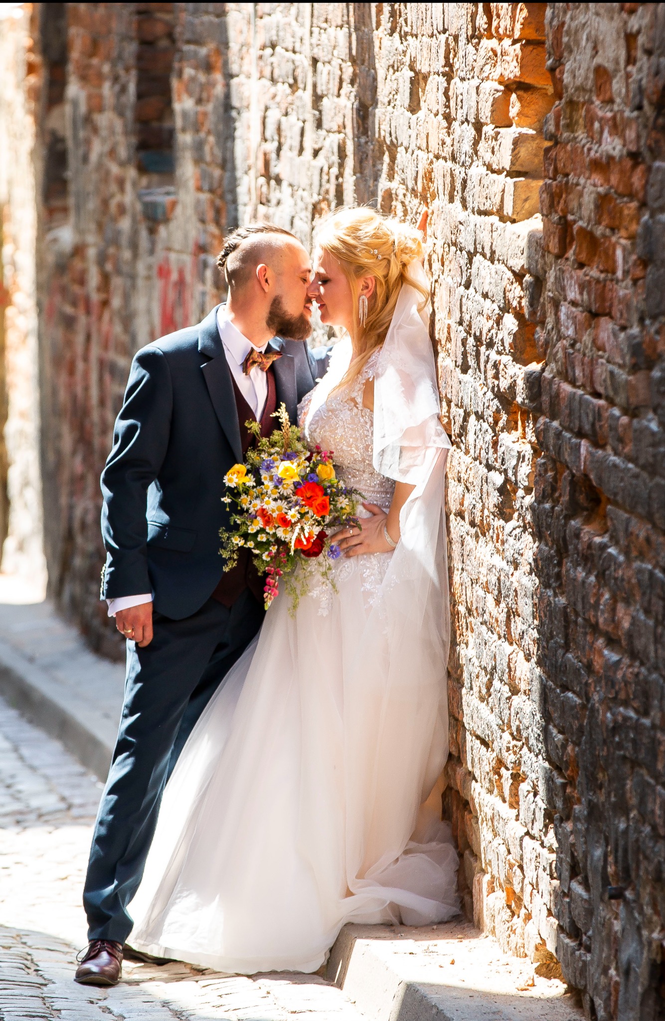 Pocałunek pary młodej w wąskiej uliczce z ceglanymi ścianami, ona w białej sukni z bukietem, on w granatowym garniturze z muszką w kwiaty, słoneczny dzień.