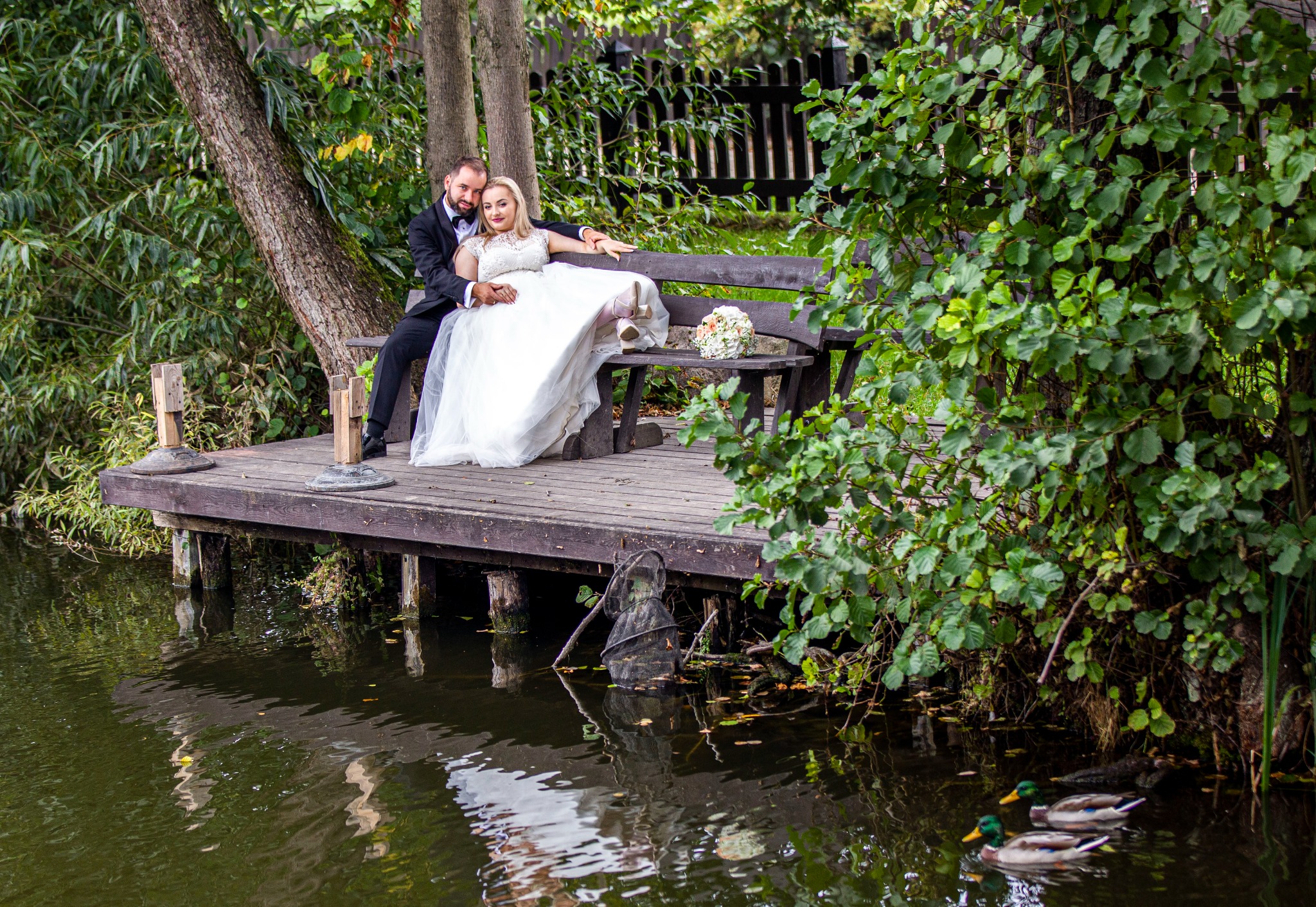 Para młoda siedzi na drewnianej ławce nad wodą w otoczeniu zieleni. Bukiet ślubny leży obok. W tle drzewa i woda z kaczkami.