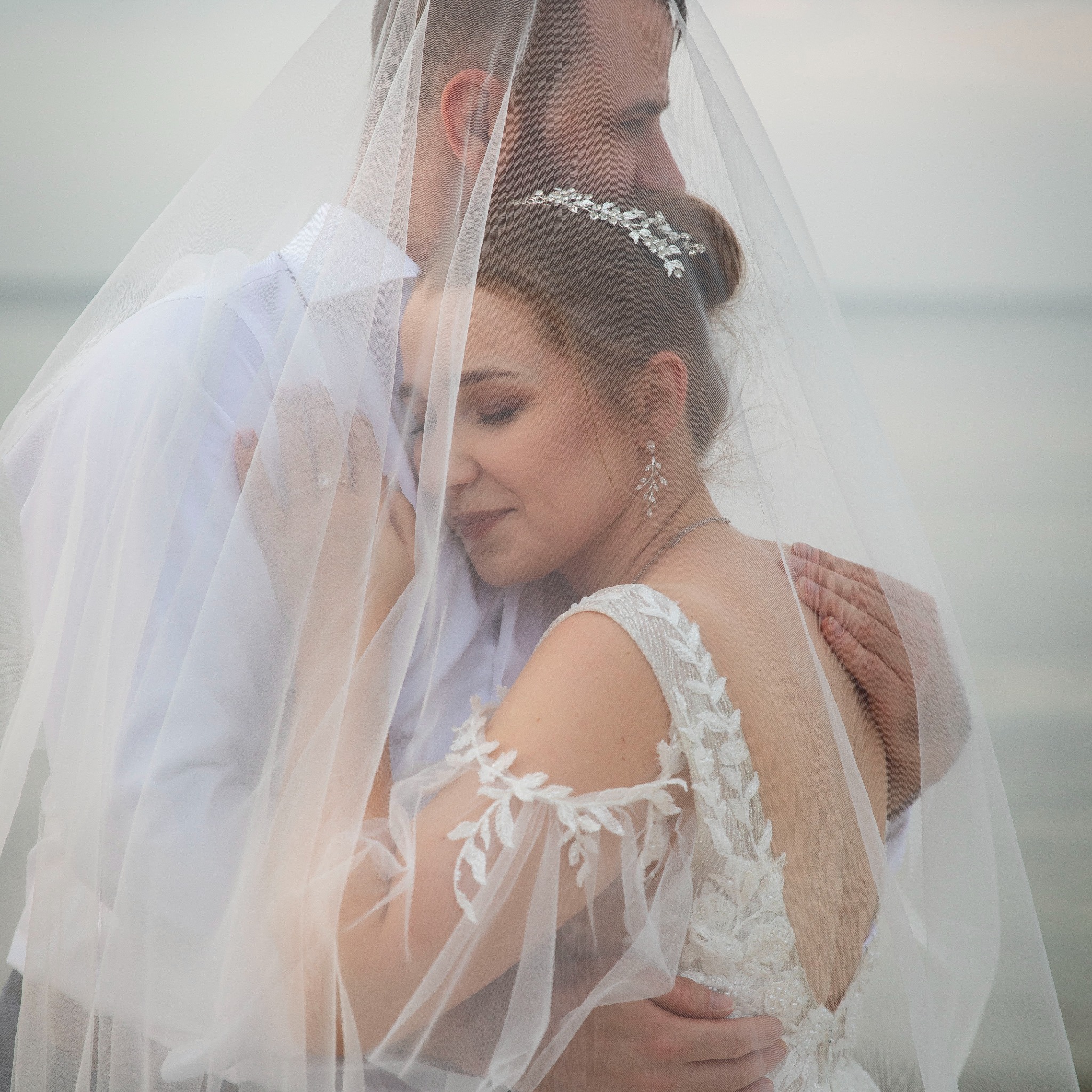 Ujęcie pary młodej otulonej welonem na tle spokojnej wody. Panna młoda w zdobnej sukni, z delikatną biżuterią, wtulona w mężczyznę w białej koszuli. Romantyczna atmosfera.