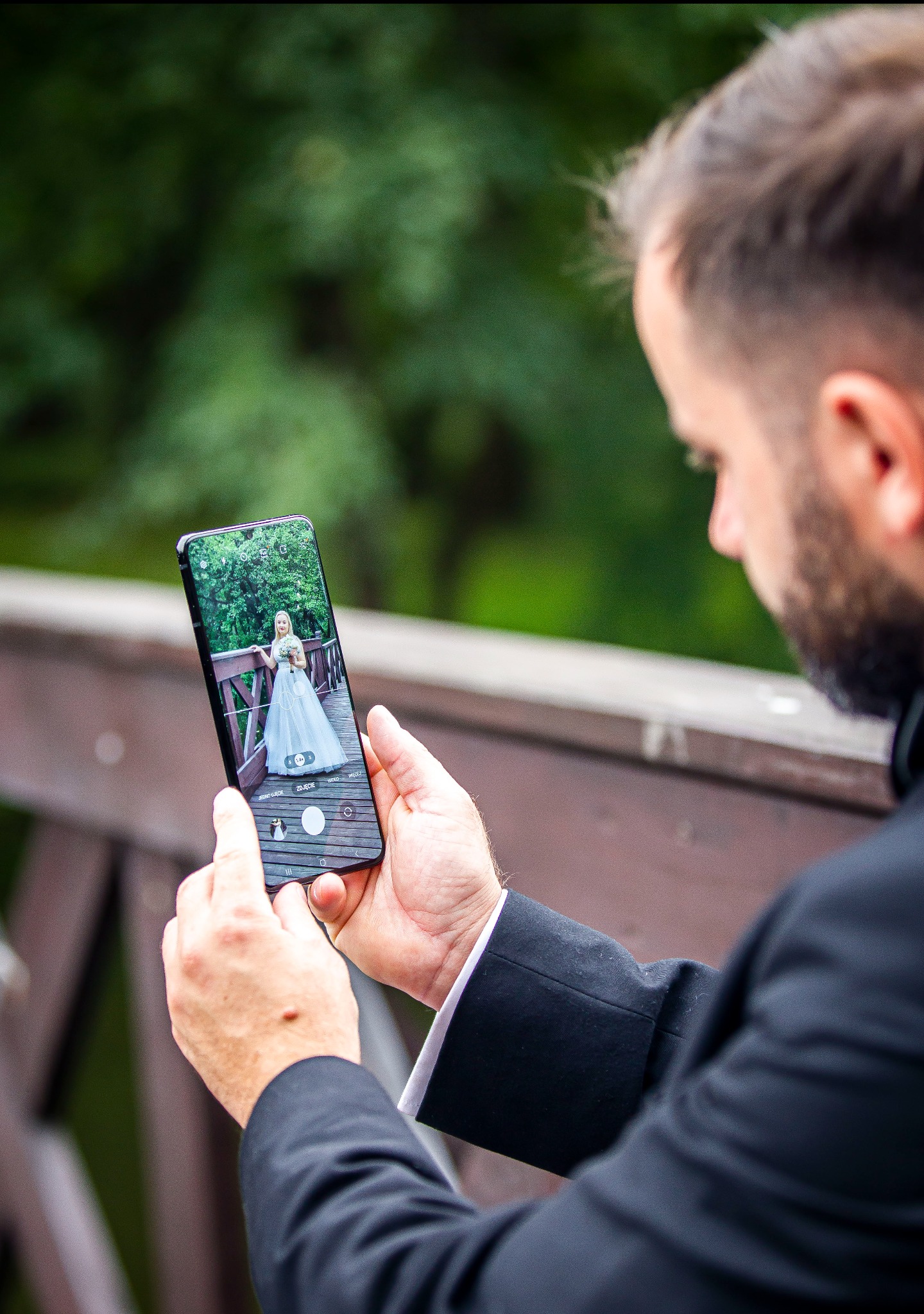 Mężczyzna w garniturze robi telefonem zdjęcie panny młodej w plenerze na drewnianym moście, z zielonym tłem. Widok zza pleców fotografa.