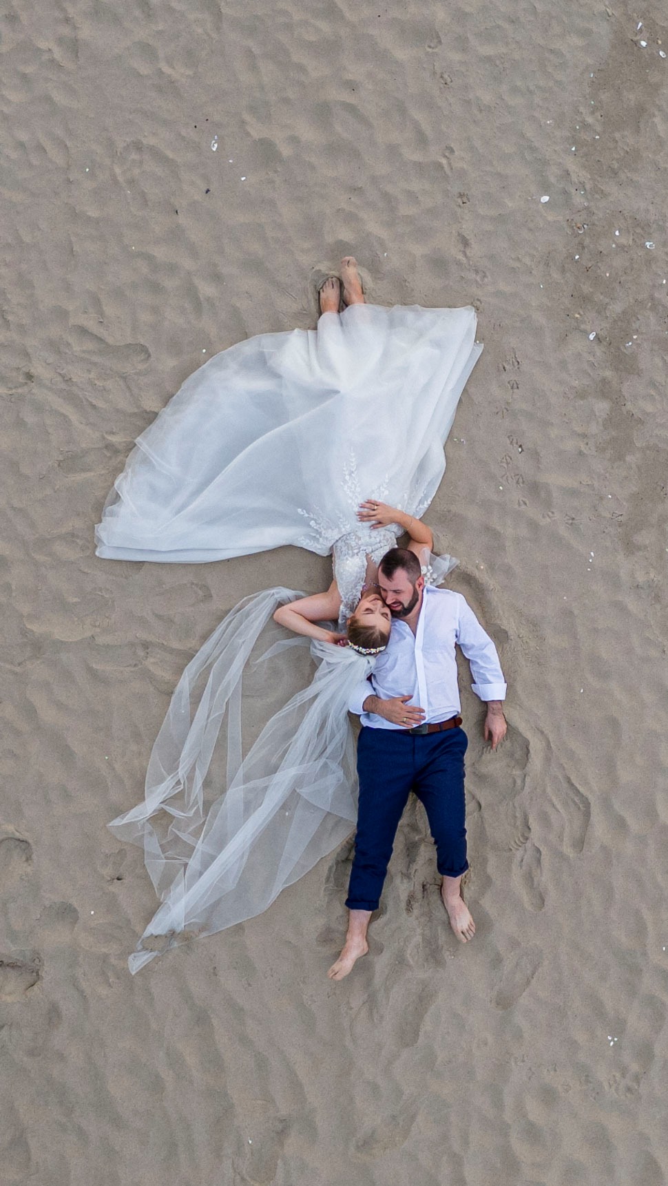 Para młoda leżąca na piasku, ujęcie z lotu ptaka. Panna młoda w białej sukni, pan młody w niebieskich spodniach i białej koszuli. Romantyczna sesja ślubna na plaży.