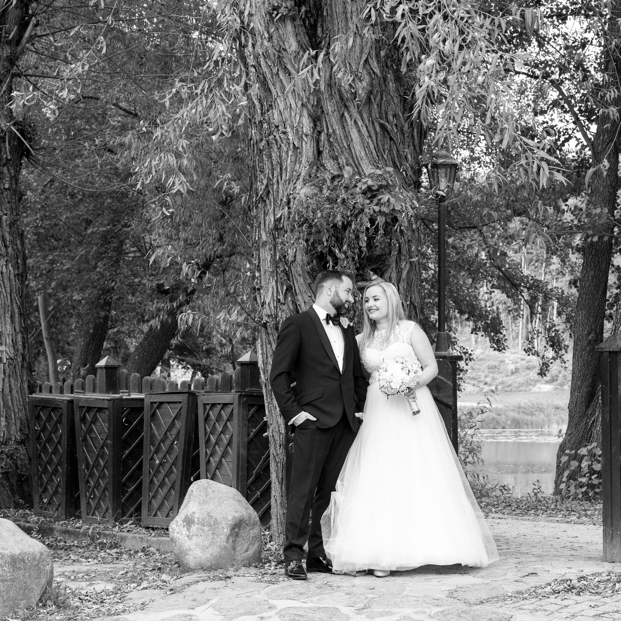 Czarno-białe zdjęcie pary młodej w eleganckich strojach, stojącej obok starego drzewa w parku, z widoczną wodą w tle i kamienną ścieżką.