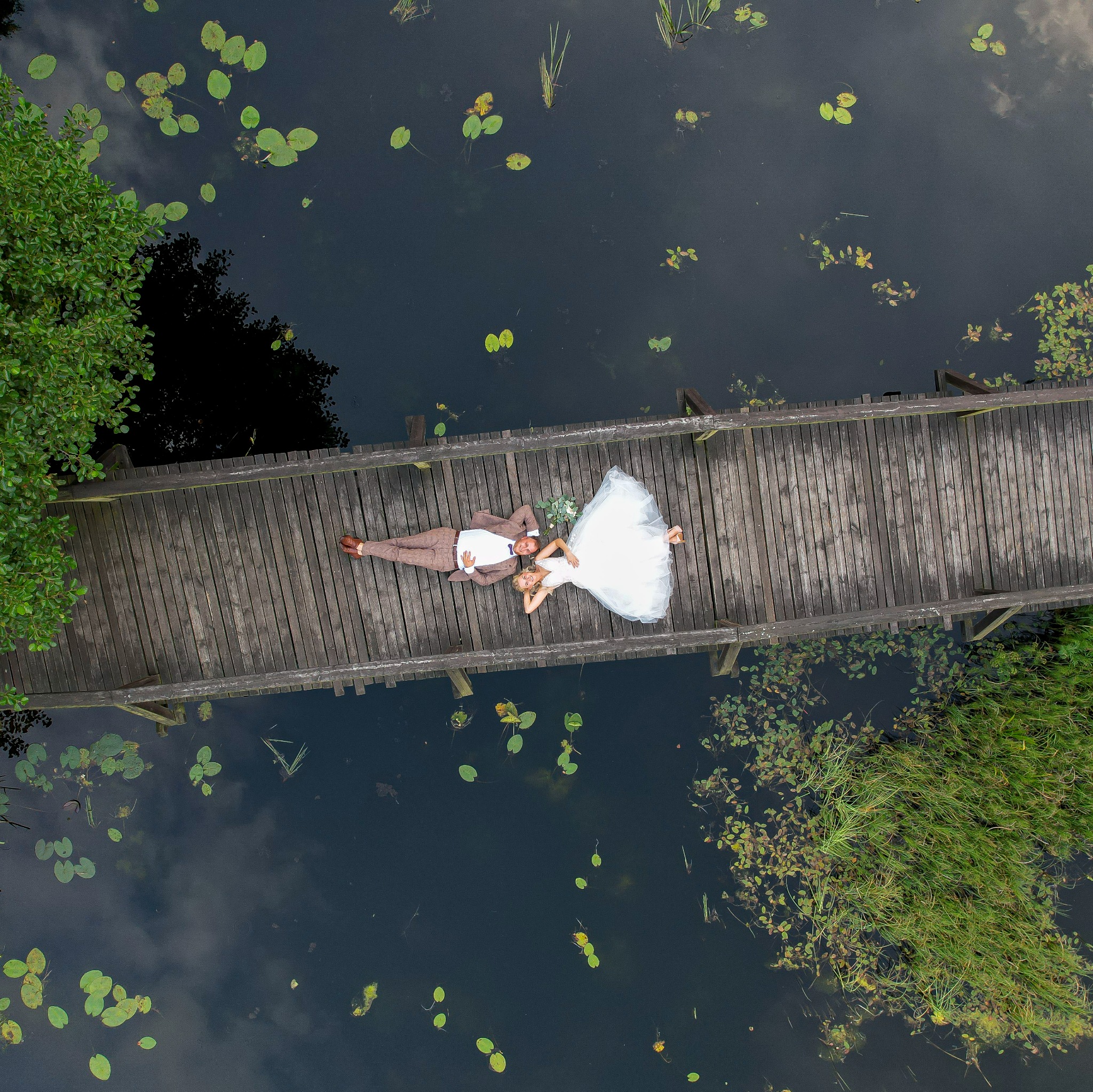 Para młoda leżąca na drewnianym moście nad stawem z liliami wodnymi, widok z góry. Pan młody w brązowym garniturze, panna młoda w białej sukni ślubnej.