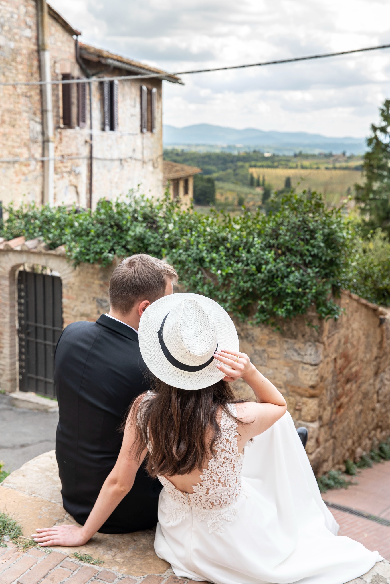 Para młoda siedzi na murku z widokiem na toskański krajobraz. Panna młoda w kapeluszu, suknia z koronkowymi plecami. Elegancki garnitur pana młodego. Kamienna zabudowa w tle.