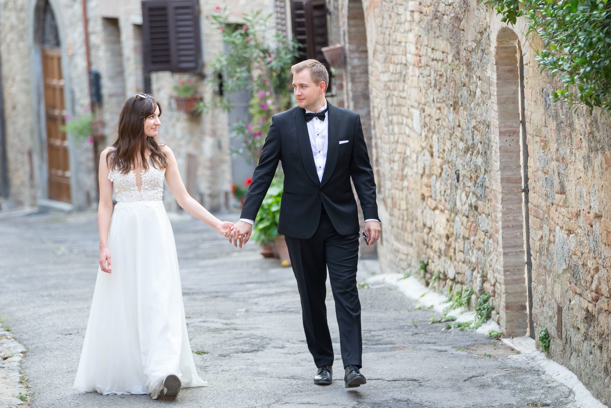 Para młoda spaceruje po malowniczej włoskiej uliczce. Ona w zwiewnej sukni, on w eleganckim smokingu. Kamienne budynki i kwiaty w tle tworzą romantyczny nastrój.