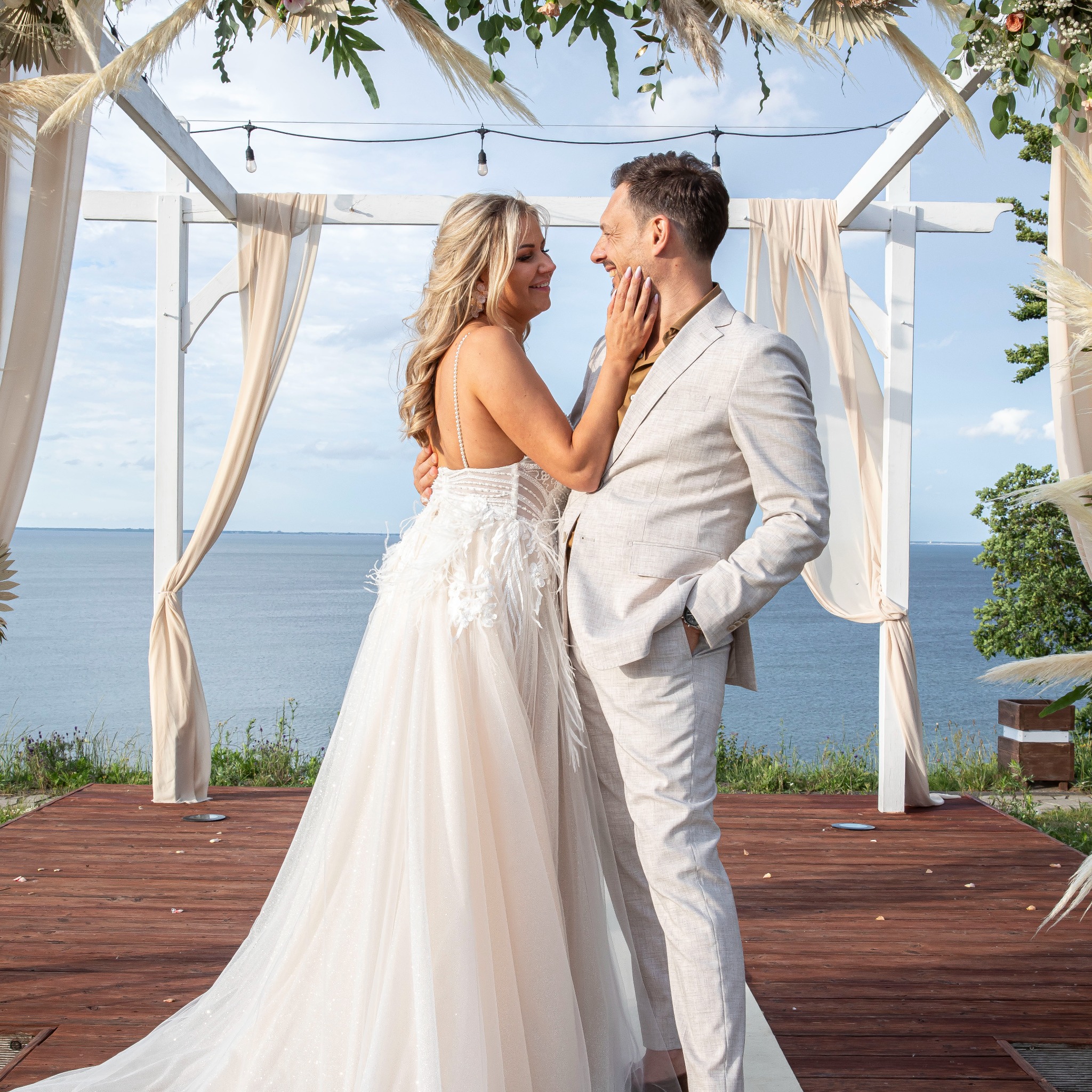 Para młoda w jasnych strojach, pod dekorowanym białym baldachimem z widokiem na morze. Ona dotyka jego twarzy, oboje uśmiechnięci.