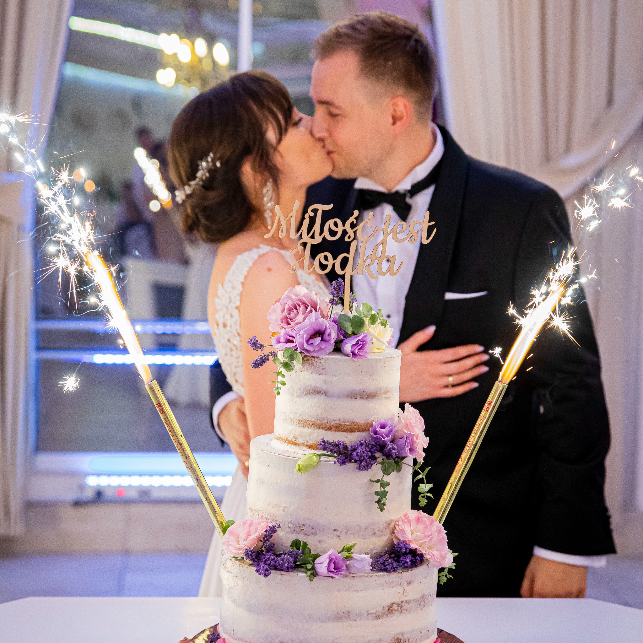 Pocałunek pary młodej za tortem weselnym zdobionym kwiatami i fontannami iskier, z napisem 'Miłość jest słodka'.