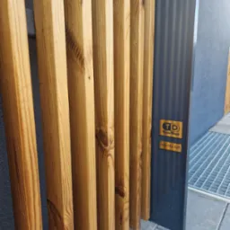 Fragment drewnianej konstrukcji z termososy 25x50mm, widoczny detal połączenia z metalowym słupkiem i logo producenta na nim, na tle szarej ściany i kratki ściekowej.