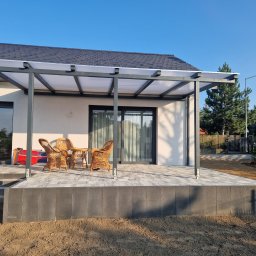 Passive House - Nowoczesny taras z szarych płyt, zadaszony konstrukcją z poliwęglanu, wyposażony w wiklinowe krzesła i stolik, widok na ogród i błękitne niebo.