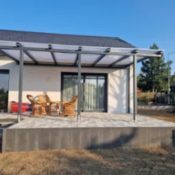 Nowoczesny taras z szarych płyt, zadaszony konstrukcją z poliwęglanu, wyposażony w wiklinowe krzesła i stolik, widok na ogród i błękitne niebo.