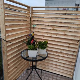 Passive House - Balkon z drewnianą przesłoną lamelową, na której tle stoi mały, okrągły stolik z dwoma doniczkami z kwiatami.