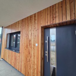 Passive House - Elewacja budynku z modrzewiową okładziną pionową, szare okno i drzwi wejściowe z długim, pionowym uchwytem.