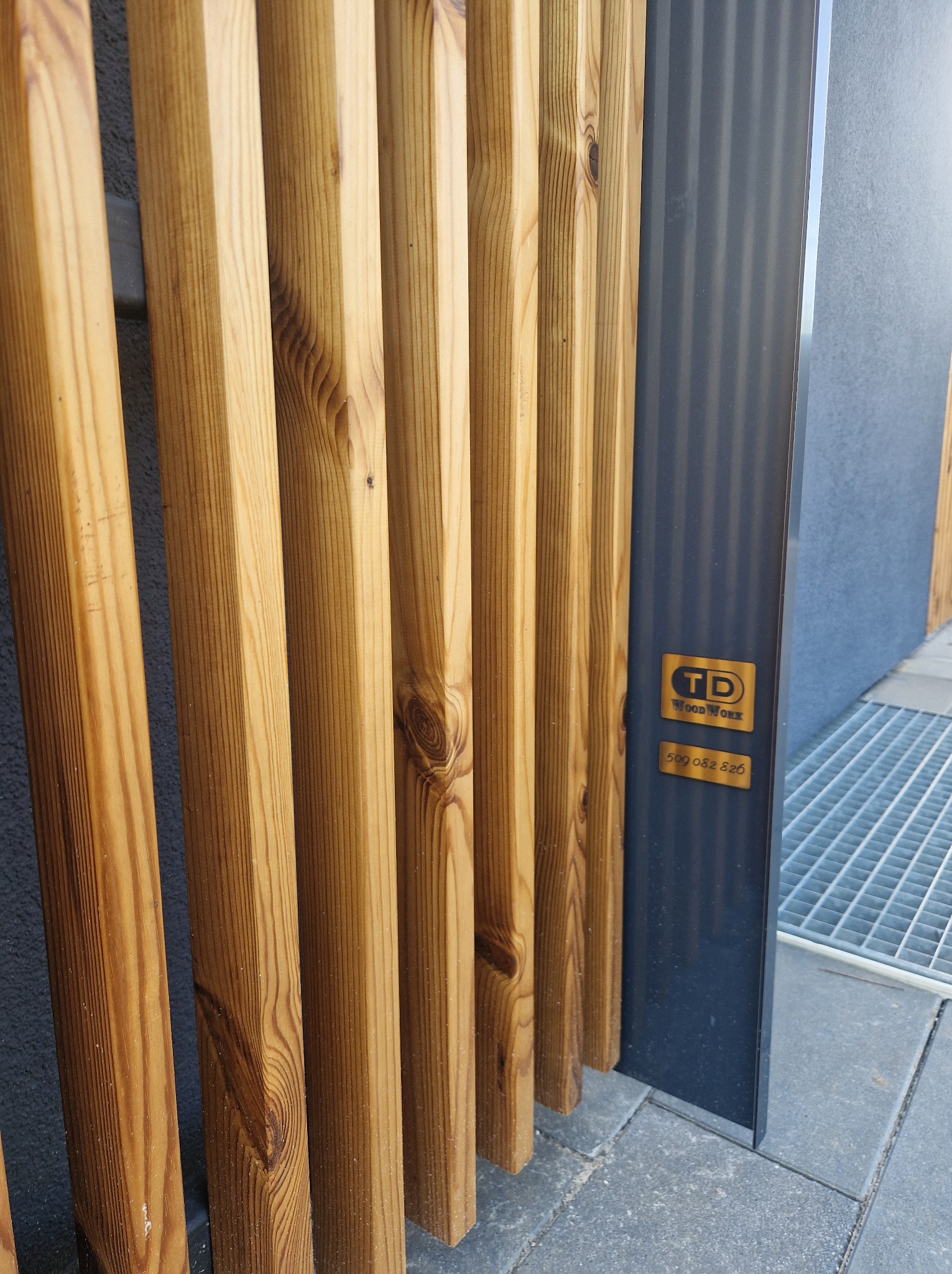 Fragment drewnianej konstrukcji z termososy 25x50mm, widoczny detal połączenia z metalowym słupkiem i logo producenta na nim, na tle szarej ściany i kratki ściekowej.